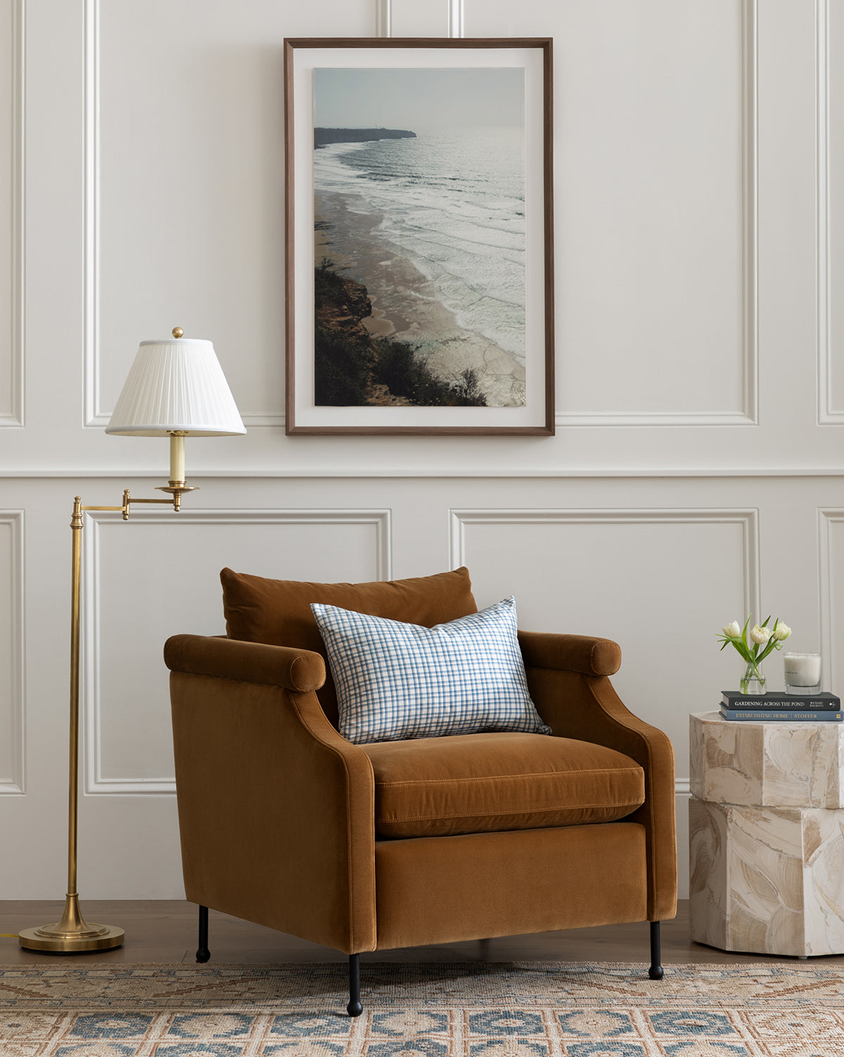 A cozy brown armchair with a plaid cushion sits next to the Dorchester Swing Arm Floor Lamp with a pleated silk shade and a small side table. A large framed ocean photograph hangs on the white paneled wall behind, by McGee & Co.