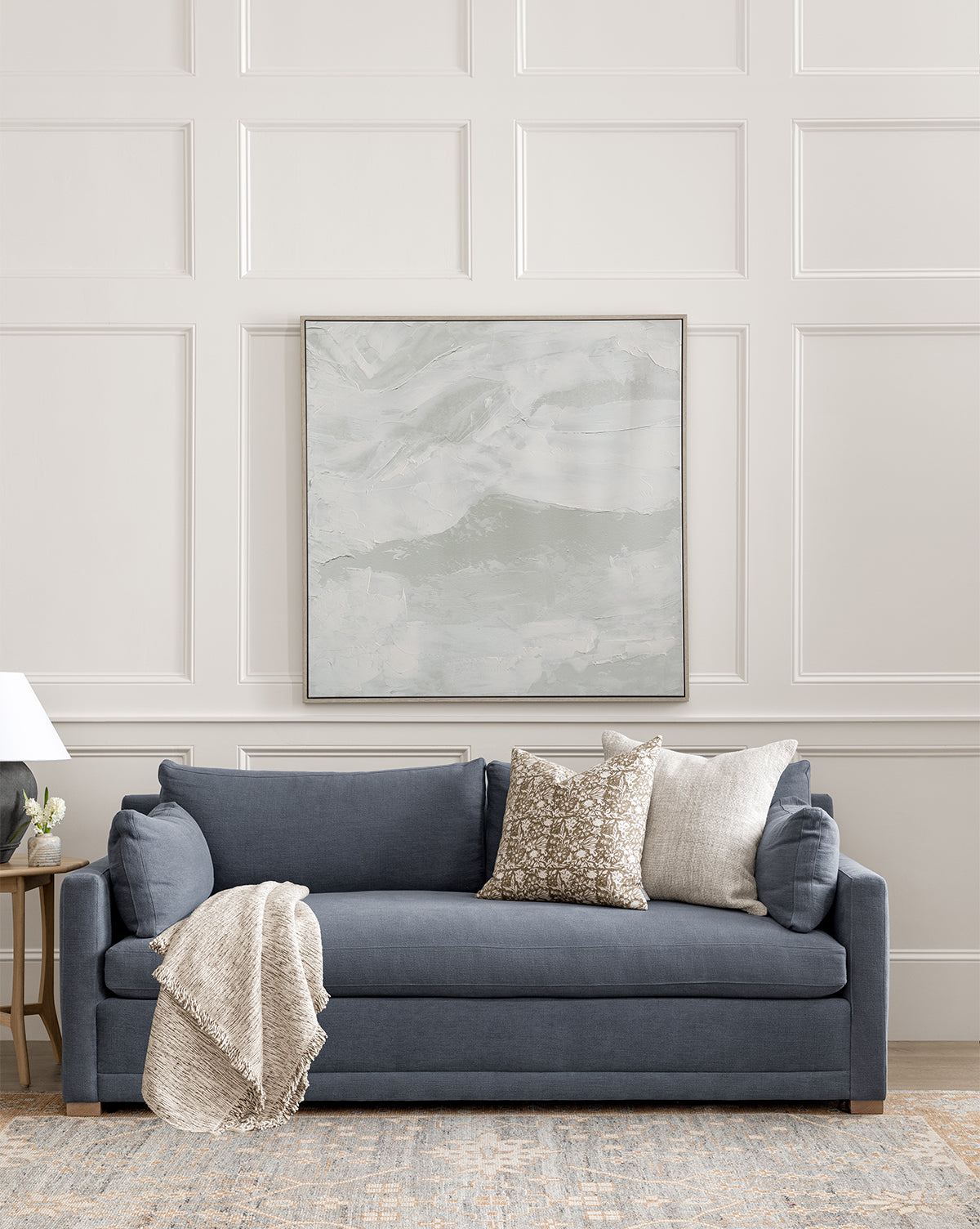 A modern living room featuring the Peterson Classic Upholstered Sofa (75 - 100) in blue, adorned with patterned and solid plush cushions and a beige throw. Above, a large abstract painting hangs on the paneled white wall with a table lamp on the left, by McGee & Co.