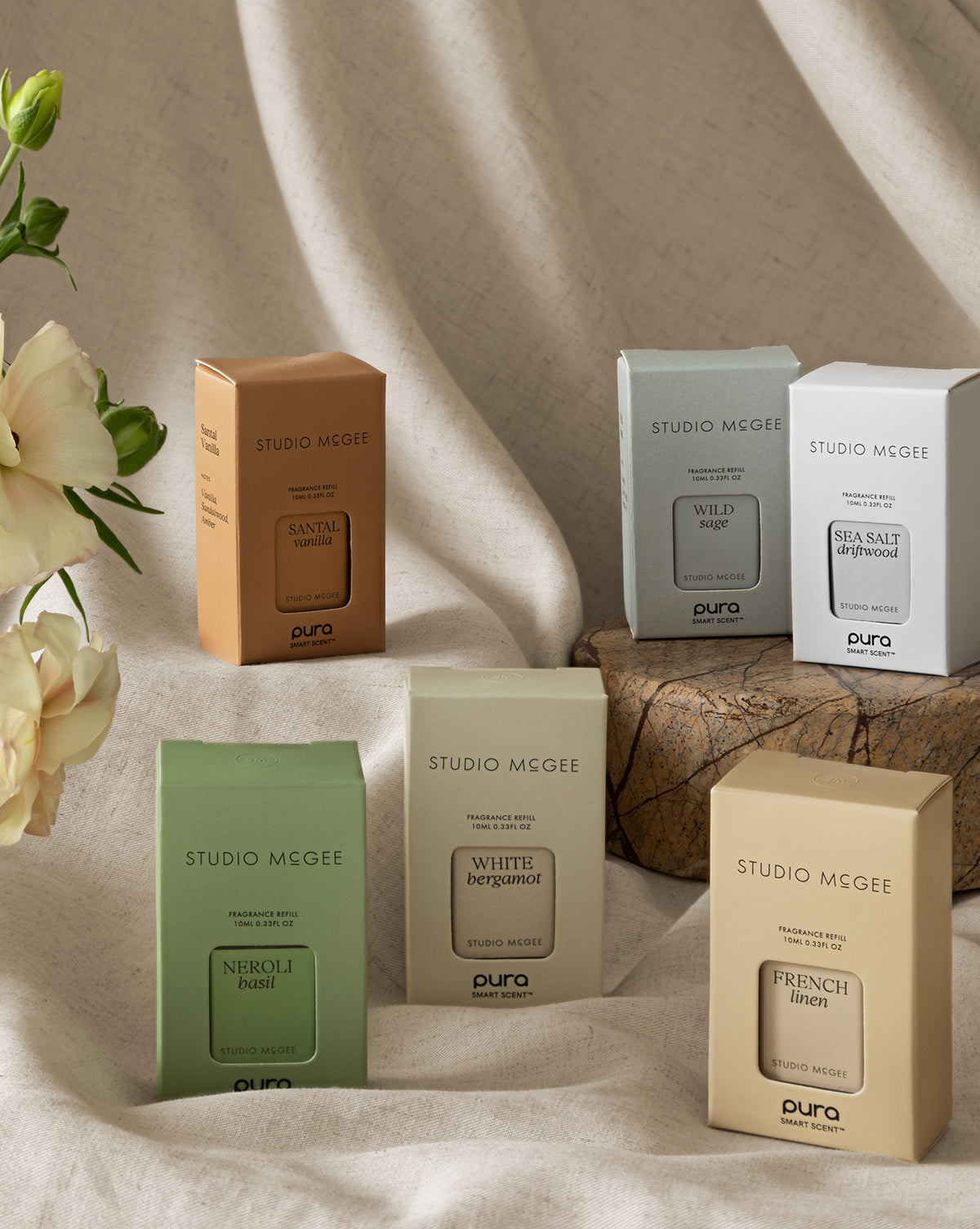 Six Pura x Studio McGee French Linen Home Fragrance Refills are arranged on a neutral fabric background with a wooden block and white flowers. Discover more scents like Santal Vanilla, Sea Salt Grapefruit, and Neroli Basil.