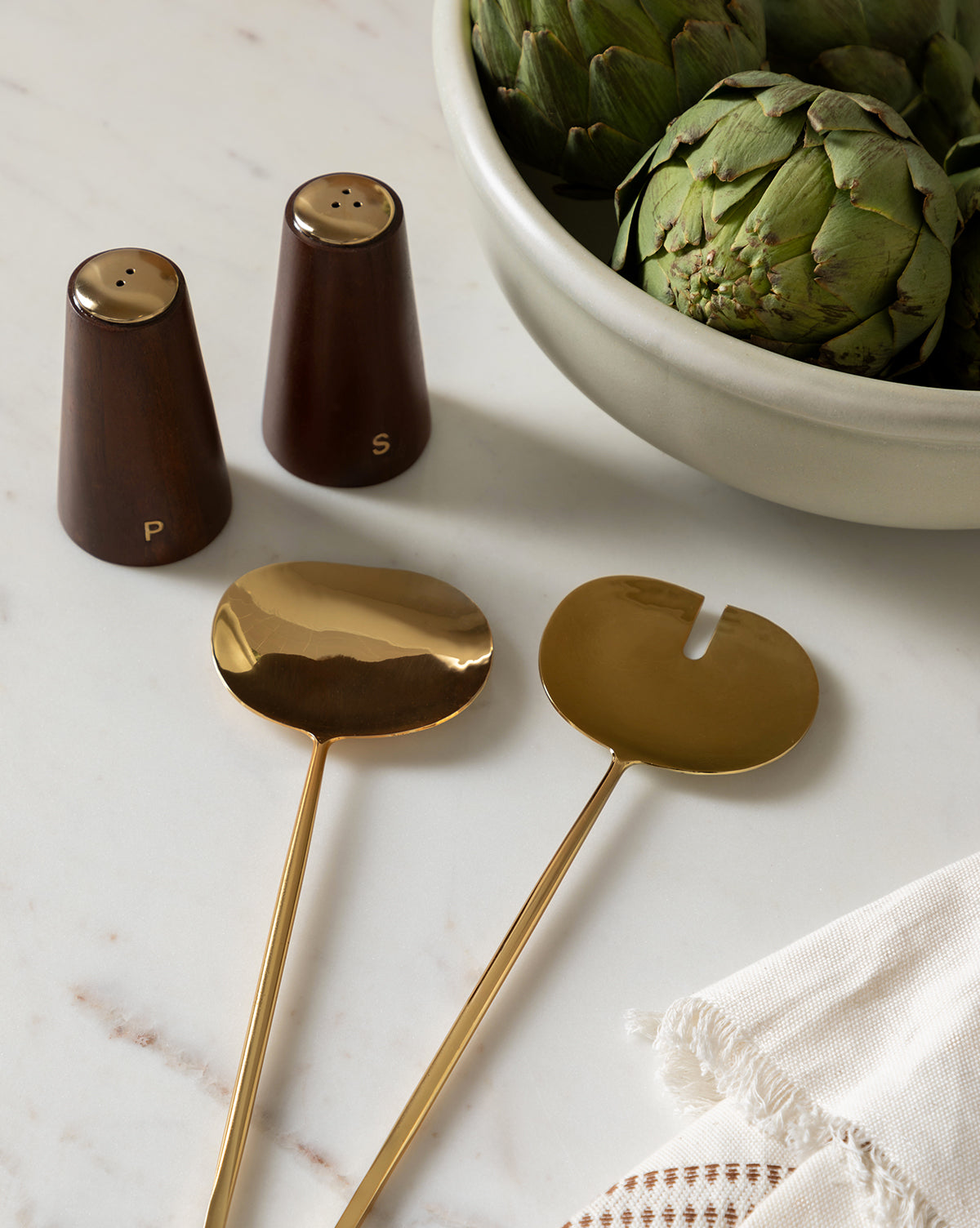 Two golden salad servers rest on a marble surface next to a beige napkin, Brasserie Salt & Pepper Shakers (Set of 2), and a bowl of artichokes—charming kitchen decor with a French café feel.