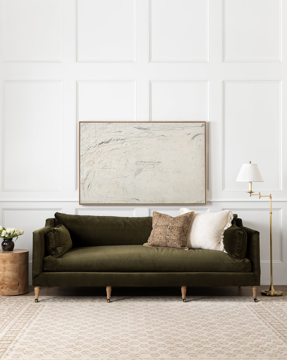 A dark green velvet sofa with neutral pillows sits in front of a white paneled wall, featuring Examination of the Rose by Tyler Guinn. A round wood side table with flowers and a gold floor lamp are beside the sofa on a patterned rug.