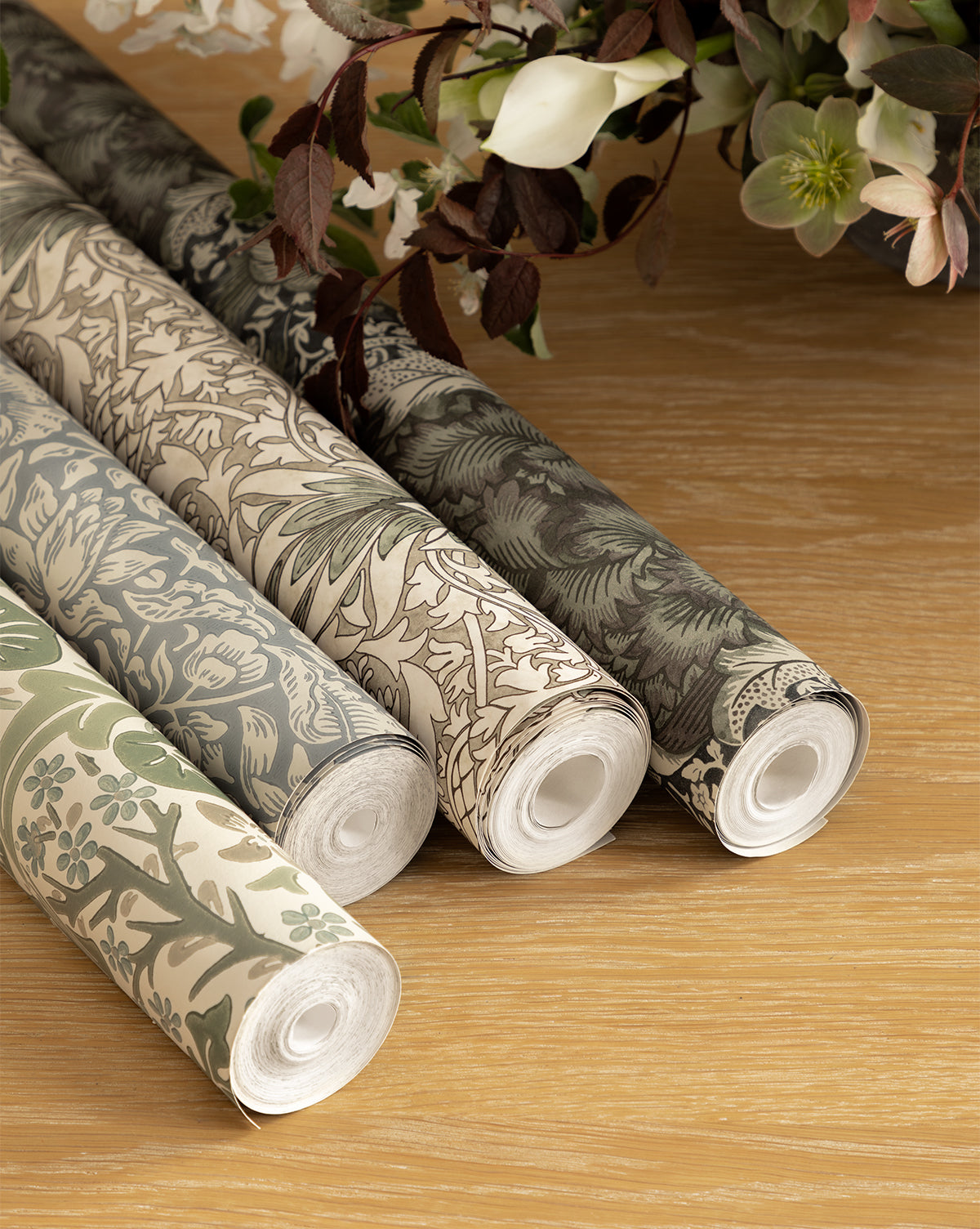 Certainly! Here is your sentence with the exact string McGee & Co. blended at the end: Four rolls of Morris & Co. x McGee & Co. Blackthorn Wallpaper by Sanderson Design Group, Inc. showcase botanical patterns in muted green, beige, and grey on a wooden surface, with white flowers and green foliage in the background, McGee & Co.