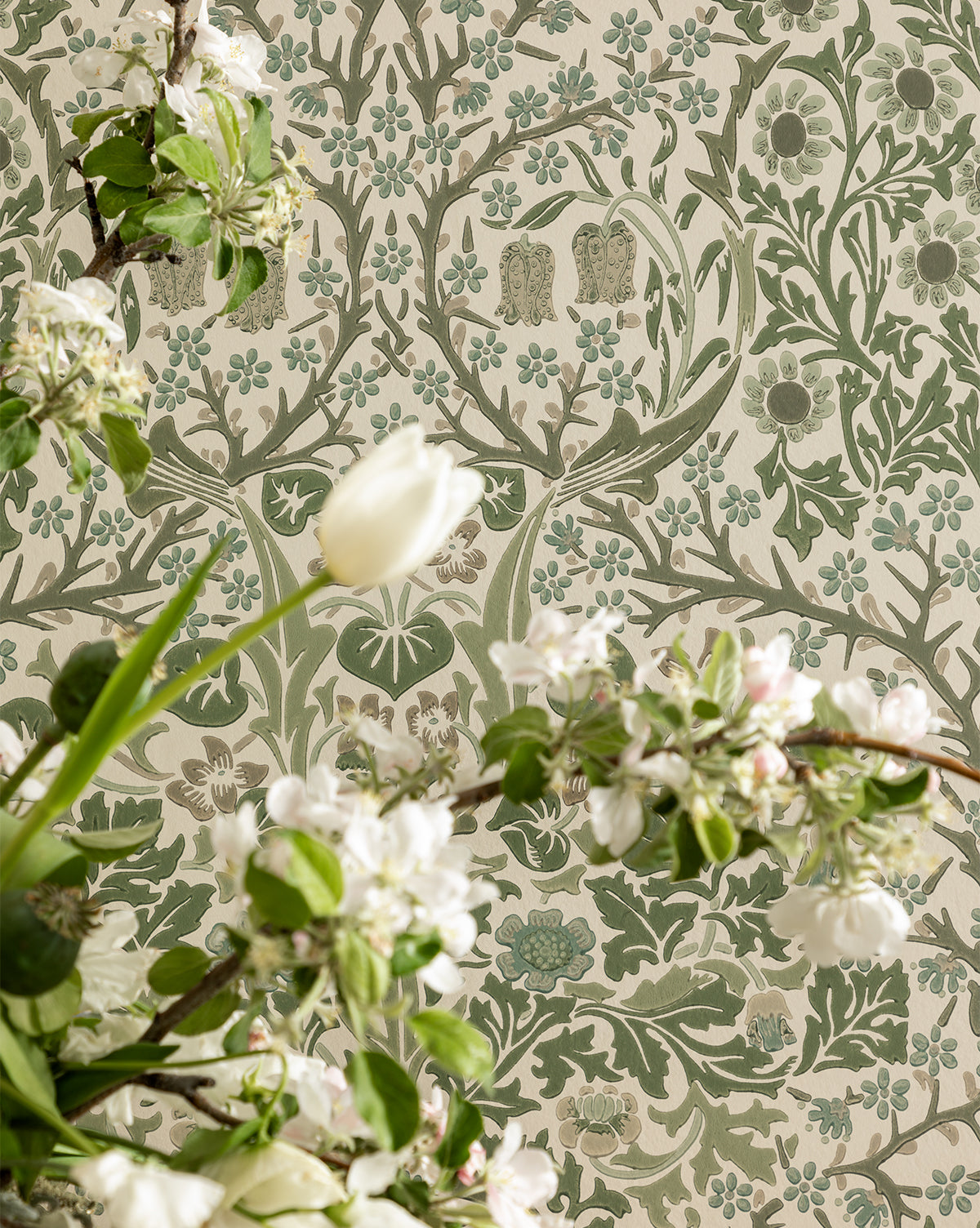 Flowering branches with white blossoms and green leaves stand before Sanderson Design Group, Inc.s Morris & Co. x McGee & Co. Blackthorn Wallpaper, which features an intricate floral pattern in muted green and beige tones, McGee & Co.