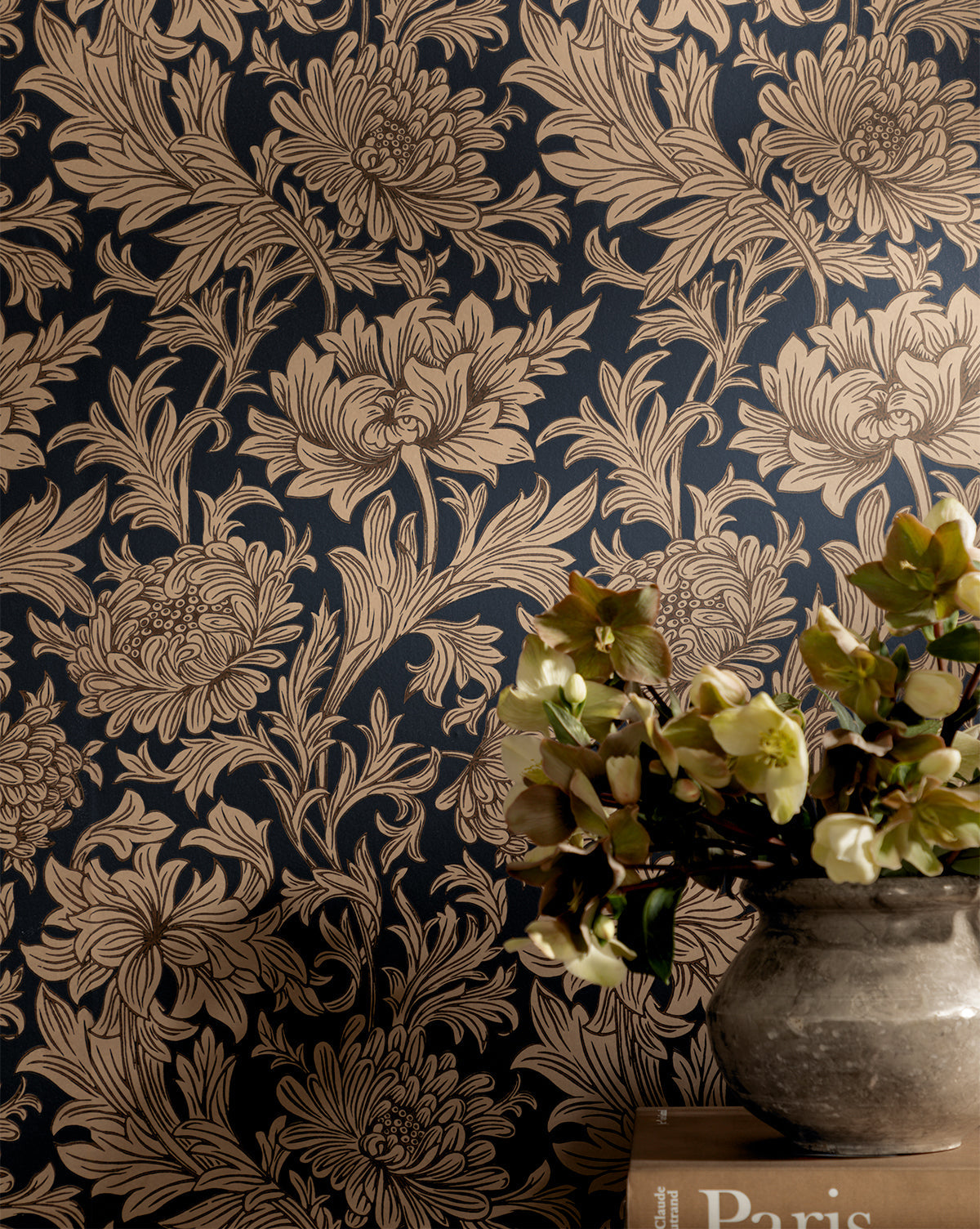 The Morris & Co. x McGee & Co. Chrysanthemum Wallpaper by Sanderson Design Group, Inc. features bold beige floral motifs on a dark background, evoking classic style. A vase with pale flowers rests on books in the lower right corner of the scene—McGee & Co.