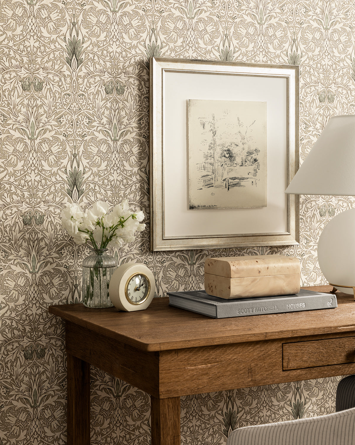 A wooden desk with a small clock, white flowers in a vase, stacked books, and a lamp sits against floral wallpaper with Etched Brunch framed above.