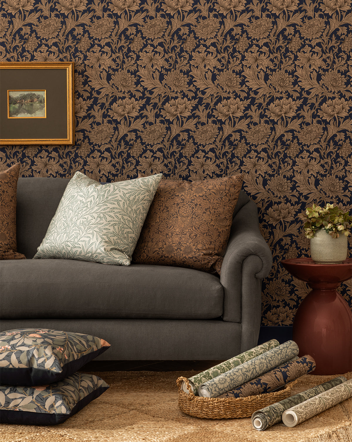 A cozy living room features a gray sofa, patterned pillows, a red side table with a plant, floral fabric accents, and a striking wall decorated with Morris & Co. x McGee & Co. Chrysanthemum Wallpaper by Sanderson Design Group, Inc., all thoughtfully curated by McGee & Co.