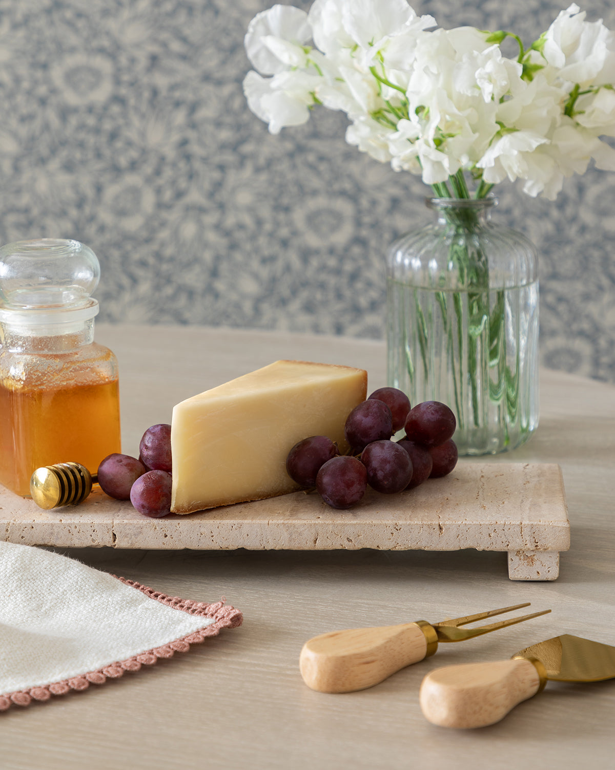 A wedge of cheese, red grapes, and a jar of honey are displayed on the Beige Travertine Footed Serving Board, with cheese knives, a napkin, and white flowers in a vase on the softly patterned tabletop, by McGee & Co.