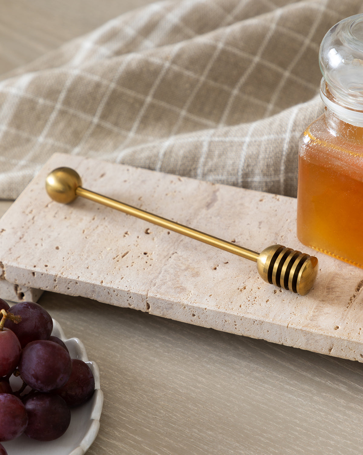 A gold honey dipper rests on a Beige Travertine Footed Serving Board beside a jar of honey, with red grapes on a white plate in the front and a checkered cloth in the background, by McGee & Co.