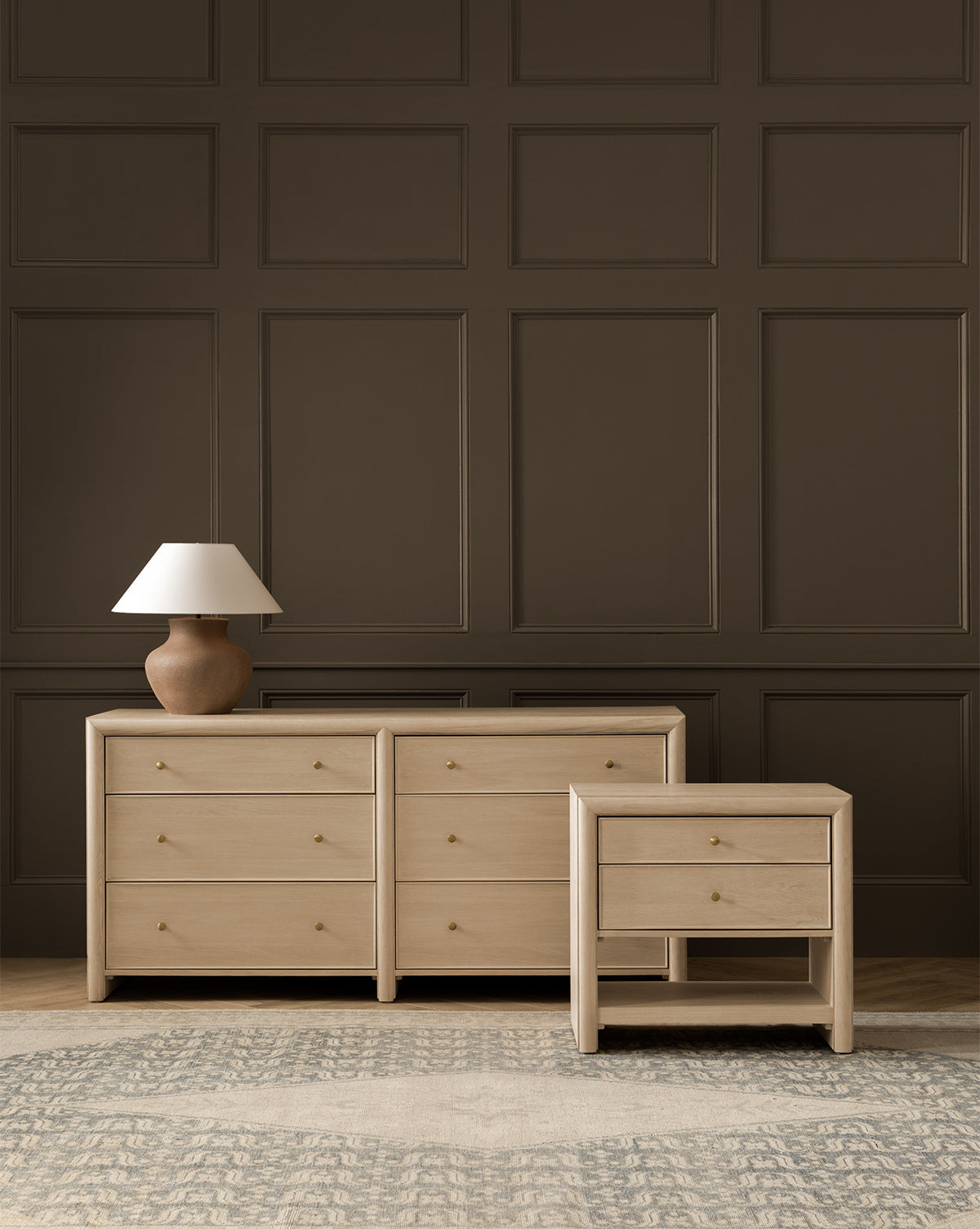 A light wood dresser with six drawers and the matching Tellerbeck Nightstand stand against a dark paneled wall, by McGee & Co. A brown ceramic lamp with a white shade rests on the dresser, while a textured rug brings warmth to the bedroom’s style, by McGee & Co.