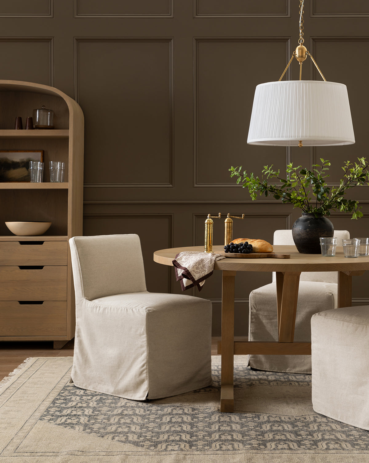 A dining room with beige slipcovered chairs, a Giselle Round Table in kiln-dried white oak, a black vase with greenery, gold utensils, a fruit plate, and a pendant light. A wooden cabinet and brown paneled walls are in the background, by McGee & Co.