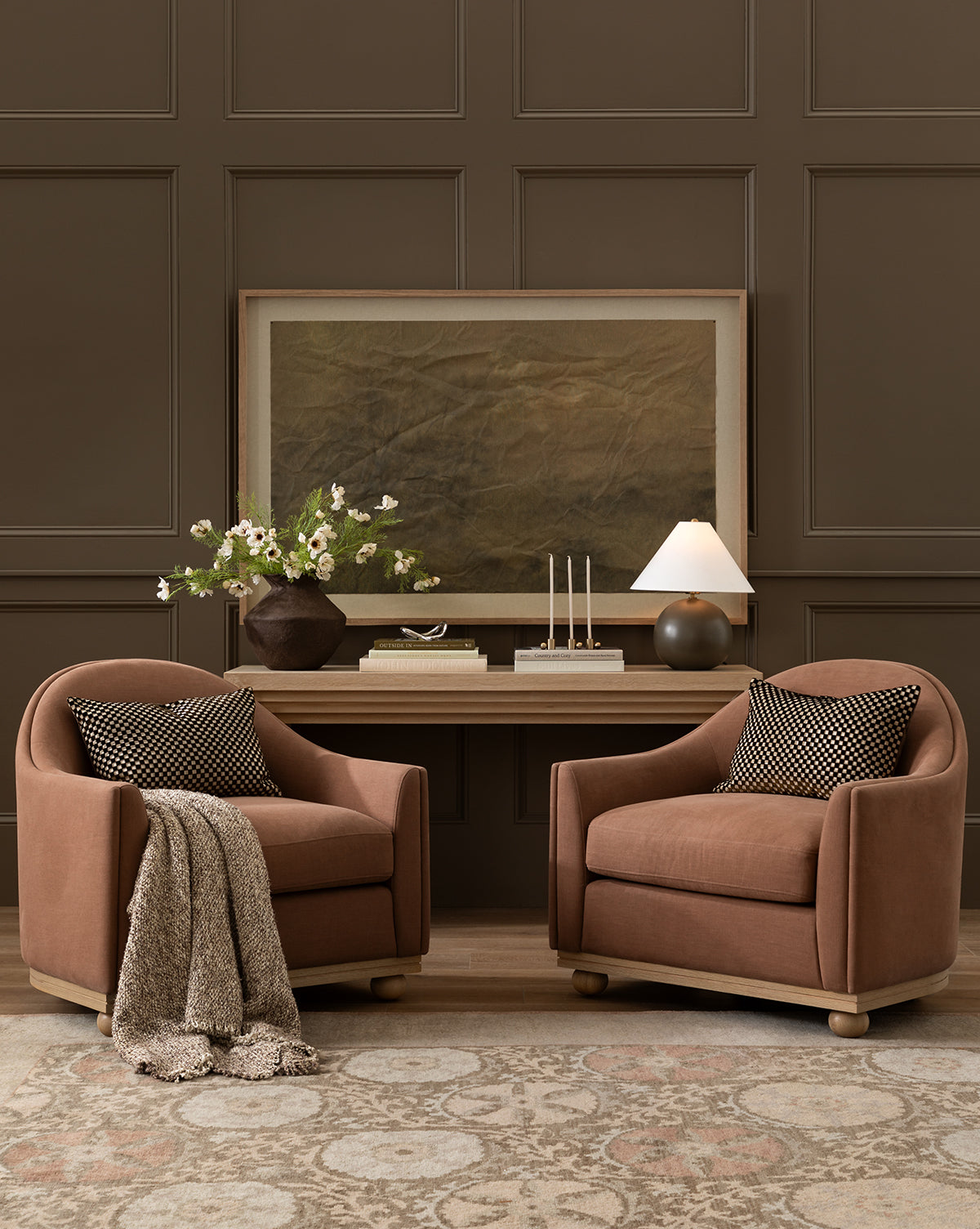 Two brown Bunnell Lounge Chairs (Ready to Ship) with patterned pillows and a textured throw are placed in front of a wooden table topped with a flower vase, candles, and a lamp, by McGee & Co. A large framed art piece hangs on the brown paneled wall behind them.