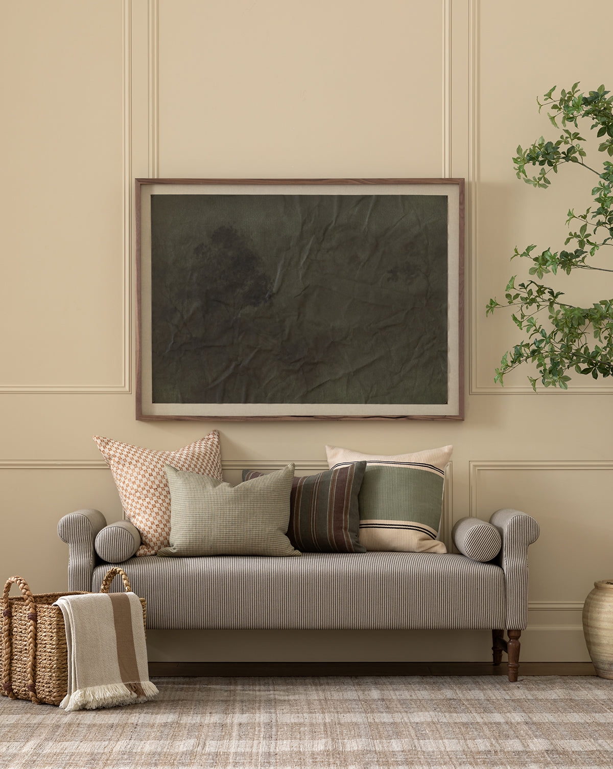 A neutral-toned living room features a light gray striped linen sofa with assorted pillows, woven baskets, a potted plant, a large dark abstract artwork above the sofa, and an elegant Asher Bench with cozy bolster pillows, by McGee & Co.