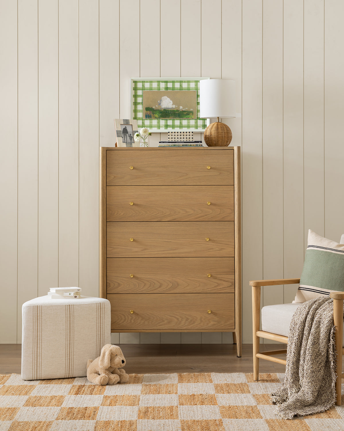 A cozy room features the Dreamscape light wood dresser with a lamp, framed art, books, and decor on top. A beige armchair with cushions, striped ottoman stacked with books, plush toy dog, and checkered rug complete the inviting space by McGee & Co.