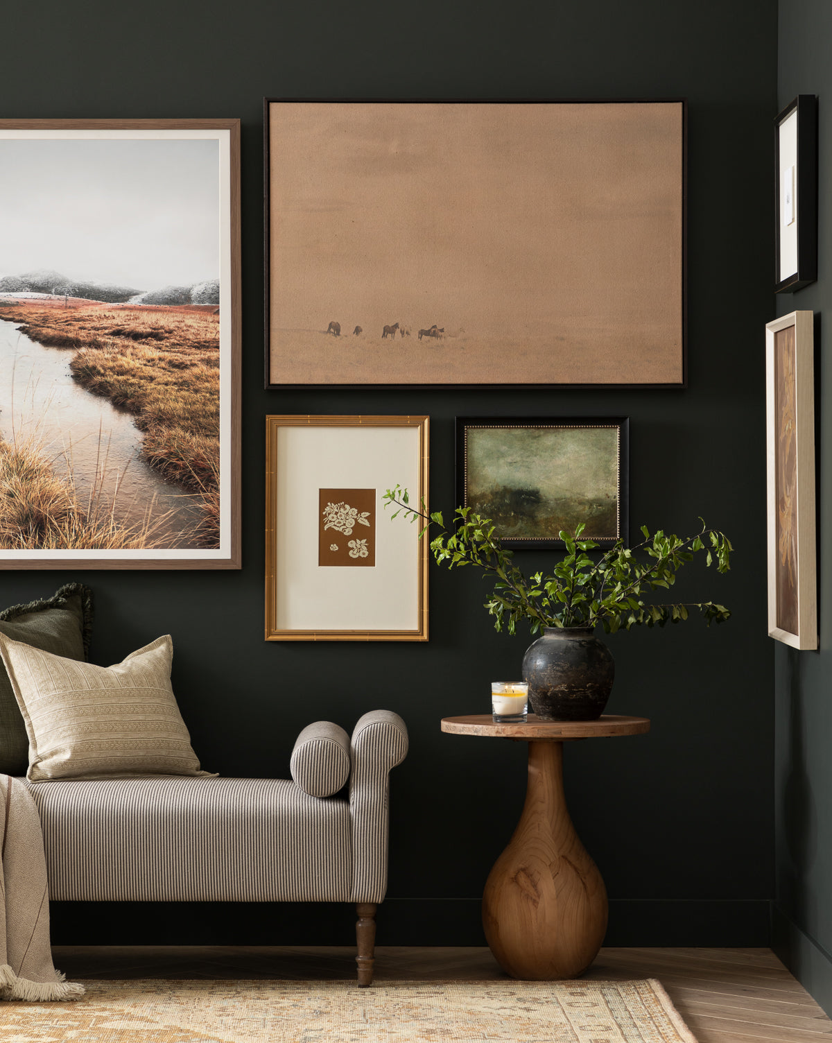 A cozy living space with a gray chaise lounge, beige pillow, round wooden side table, black vase with greenery, and the Smokey Pasture country landscape wall art framed on a dark green wall, by McGee & Co.