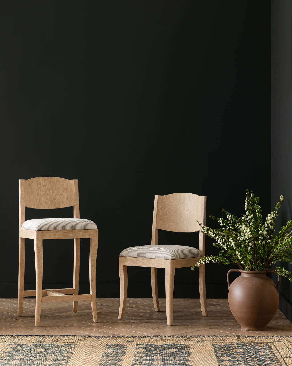 Two Shepherd Dining Chairs in light wood, featuring striped upholstered seats, are placed on a wooden floor against a black wall—one in standard height and one taller like a barstool—next to a large brown vase of green and white flowers by McGee & Co.