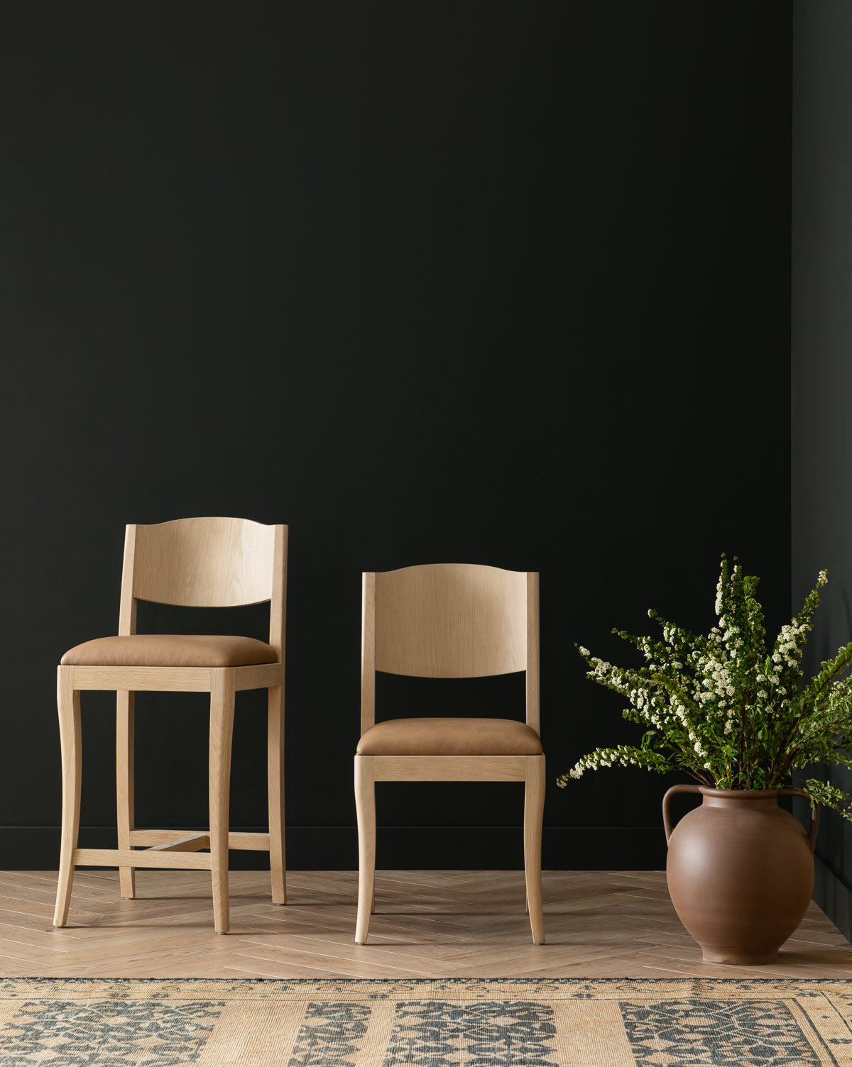 Two Shepherd Dining Chairs with white oak frames and tan upholstered seats are set on a wooden floor by a black wall. A large ceramic vase with green branches stands nearby, and a patterned rug appears in the foreground, by McGee & Co.
