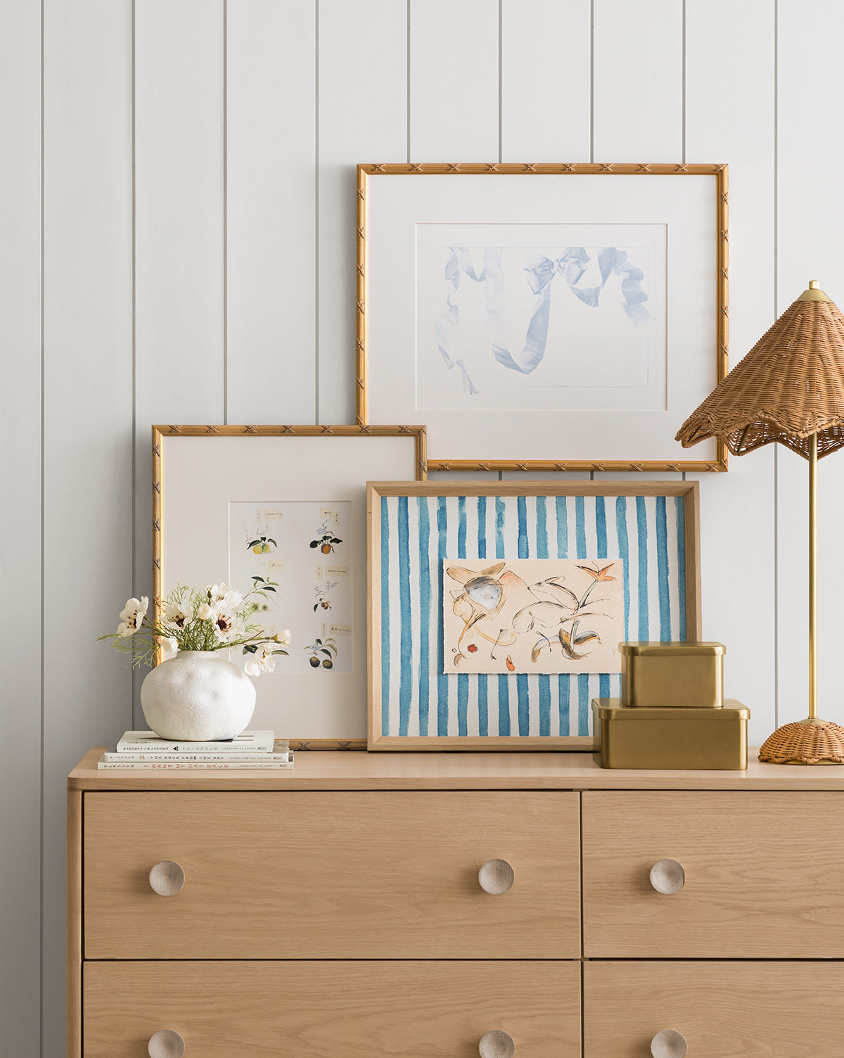 A light wooden dresser with round knobs displays decor—white vase of flowers, stacked books, gold boxes—and features three framed art prints including Leaping Doe among the Flowers, all set against a white paneled wall by McGee & Co.