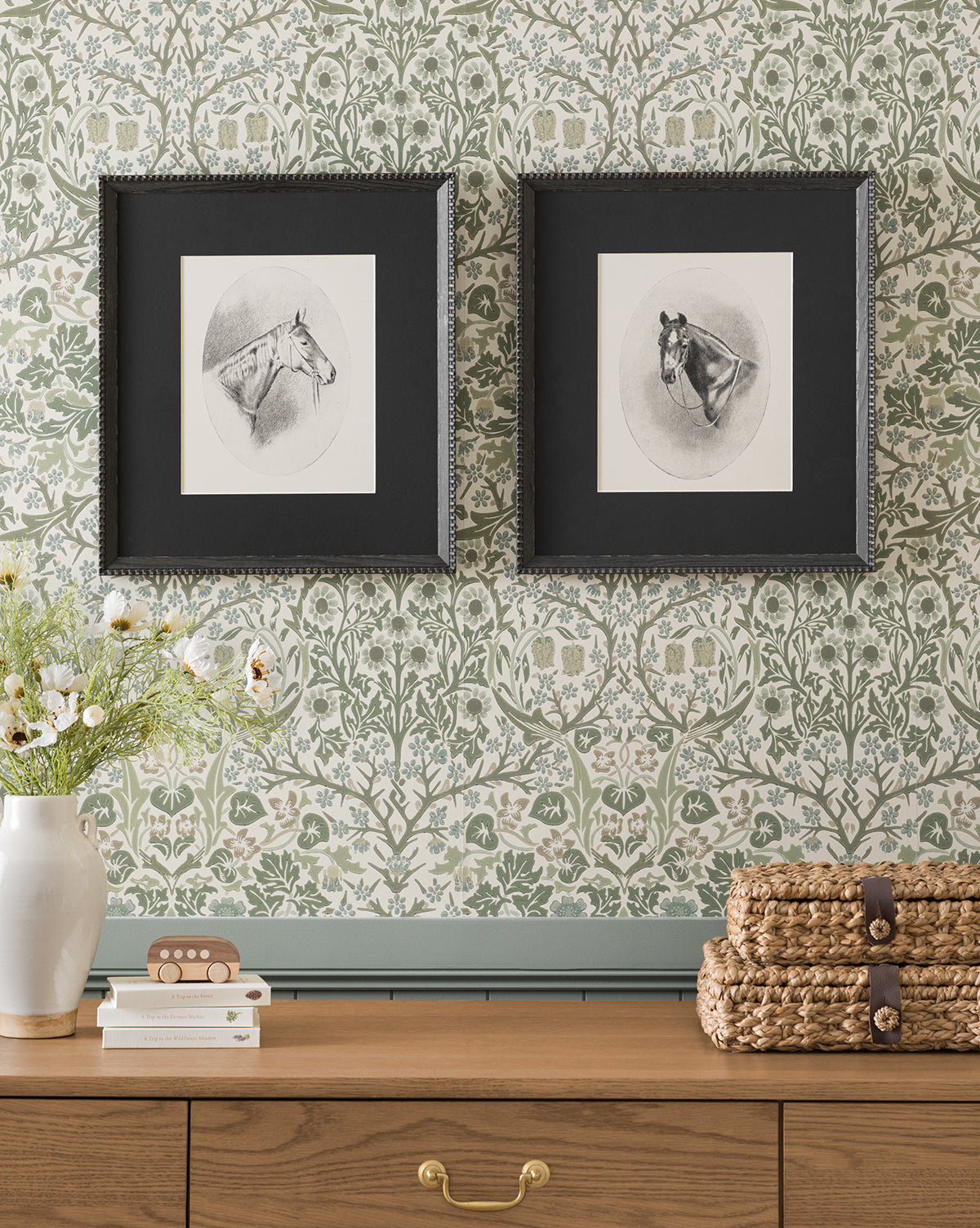 The Spirit framed horse portrait in a black frame hangs on floral wallpaper above a wooden dresser adorned with flowers, woven baskets, and decorative wall art, by McGee & Co.