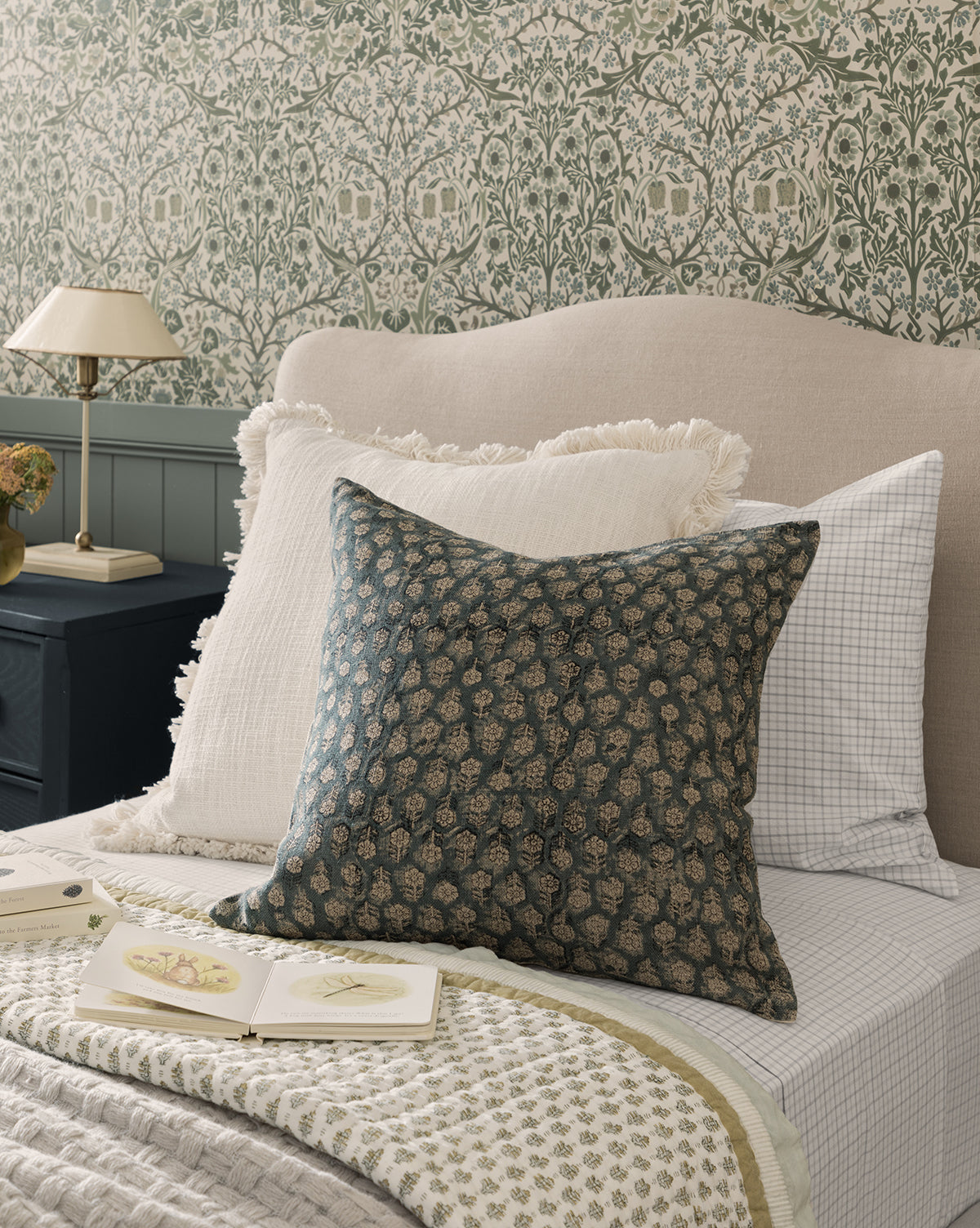 A cozy bed with layered pillows, including the Perla Pillow Cover and a hand block-printed pillow, rests against a beige upholstered headboard. Green floral wallpaper, a nightstand with flowers, and an open book complete the charming scene, by McGee & Co.