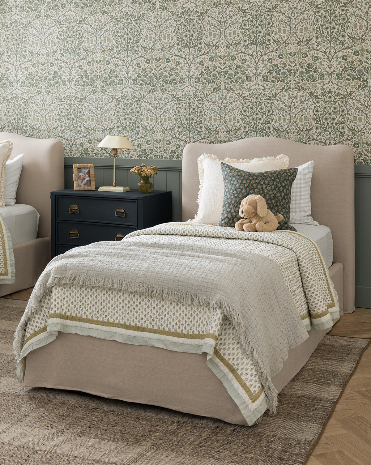 A cozy bedroom with a neatly made twin bed featuring the Dulcie Block Print Kids Quilt by Renuka Inc., beige bedding, a stuffed dog plush on the pillow, a black nightstand with a lamp and books, and green floral patterned wallpaper, finished with touches from McGee & Co.