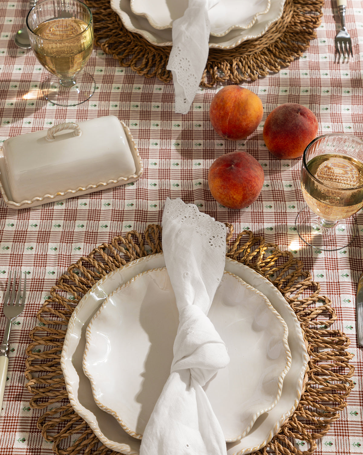 A table set for two features scalloped white plates on McGee & Co. x Loeffler Randall Elise Seagrass Placemats, white napkins, glasses of white wine, three peaches, and a ceramic butter dish atop a red-and-white checked tablecloth by McGee & Co.