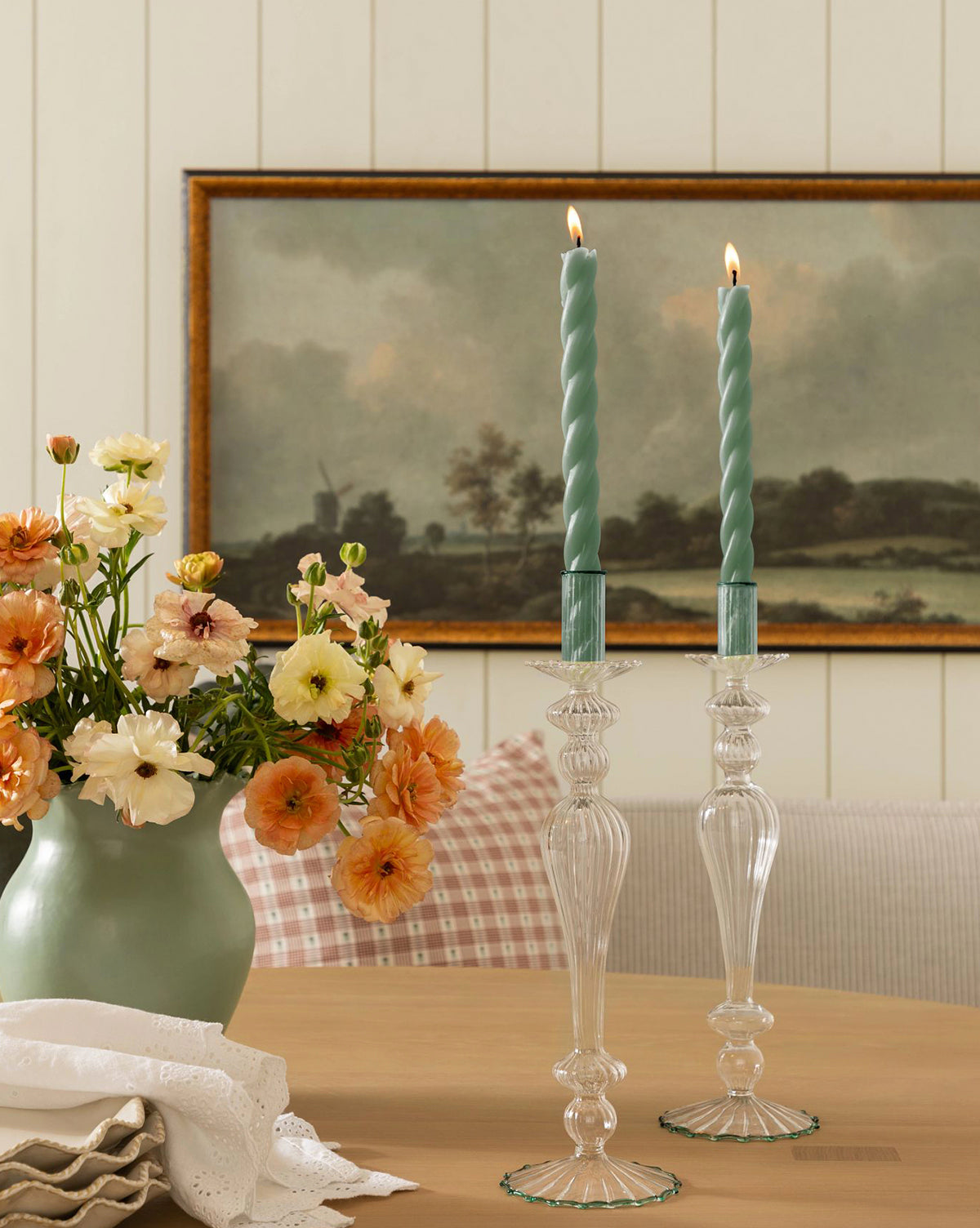 A table with a light green vase of orange and cream flowers, a set of two lit McGee & Co. x Loeffler Randall Sybil wax taper candles in glass holders, and a framed landscape painting on a paneled wall in the background by McGee & Co.
