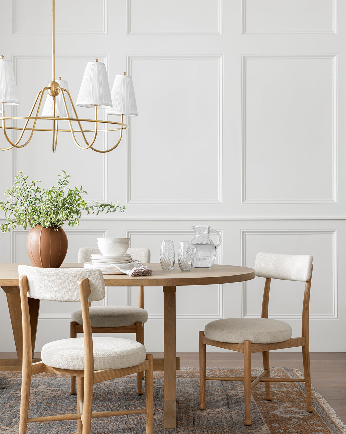 A round wooden dining table with four Garance Dining Chairs sits on a patterned rug. The table is decorated with a potted plant, dishes, glasses, and a glass pitcher. A gold chandelier hangs above against decorative white wall paneling, by McGee & Co.