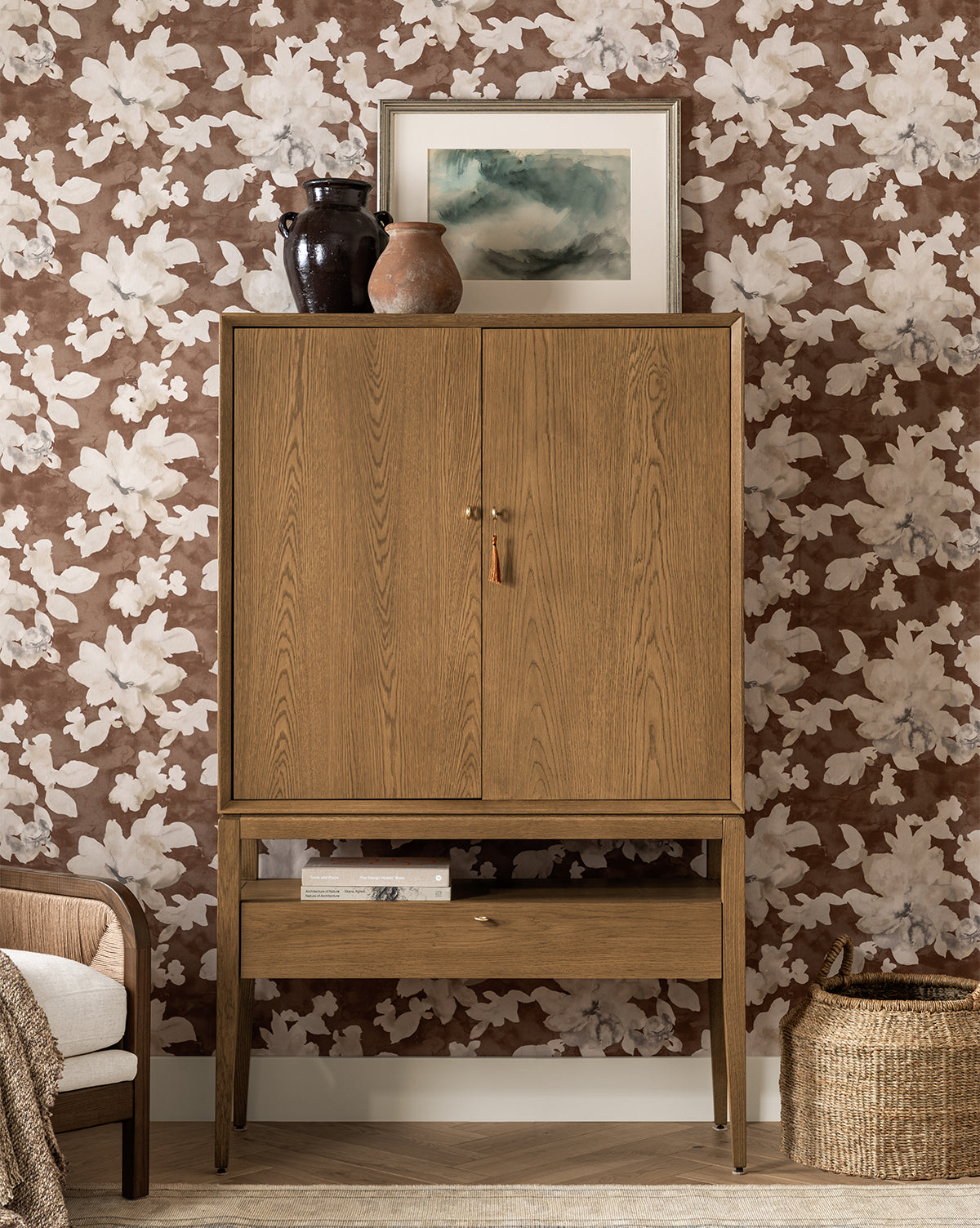 A Jahnsen Cabinet with a honey oak finish stands against floral wallpaper. Two ceramic vases and a framed abstract artwork are displayed on top, while nearby, a rattan basket and an armchair can be partially seen by McGee & Co.