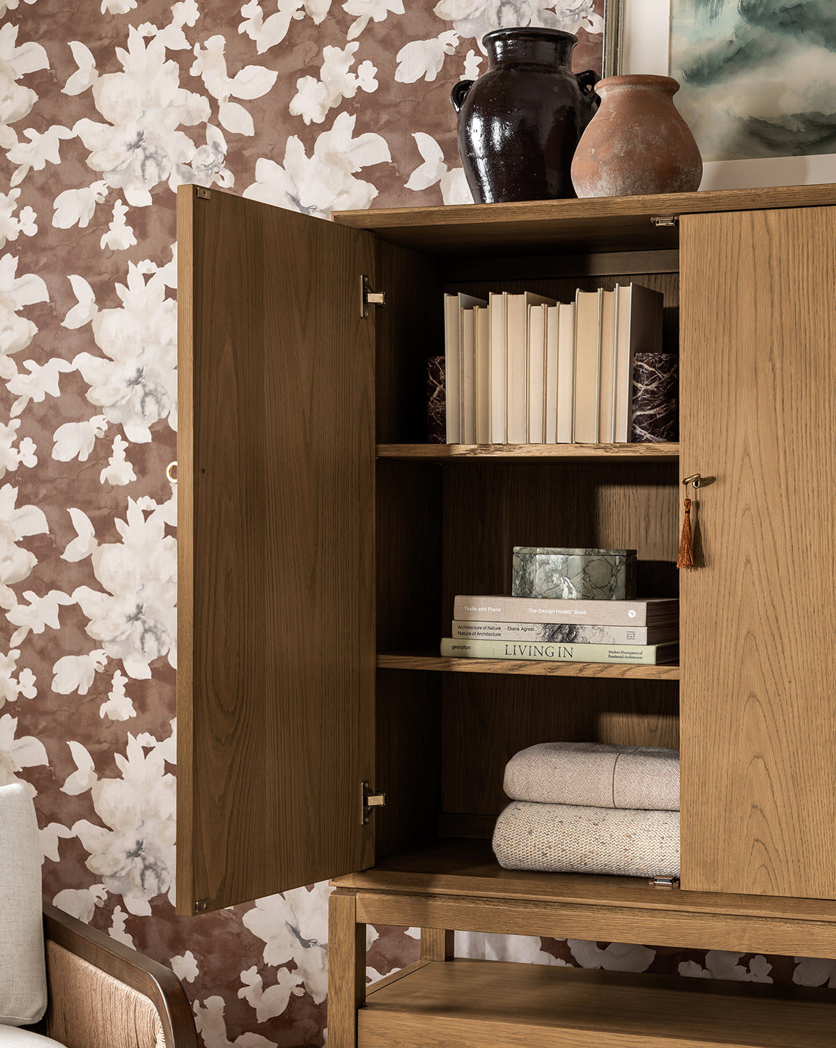 The Jahnsen Cabinet in honey oak stands with one door open, revealing books, folded textiles, a green marble box, and two clay vases. Brown floral wallpaper serves as the backdrop, by McGee & Co.