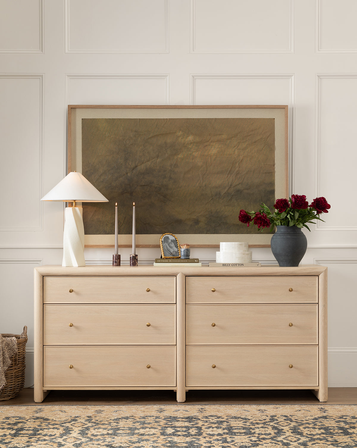 A light wood Tellerbeck Dresser with gold knobs showcases a lamp, candles, decor, and a vase of red flowers beneath large framed art. A woven basket on the floor nearby completes this stylish bedroom scene, by McGee & Co.