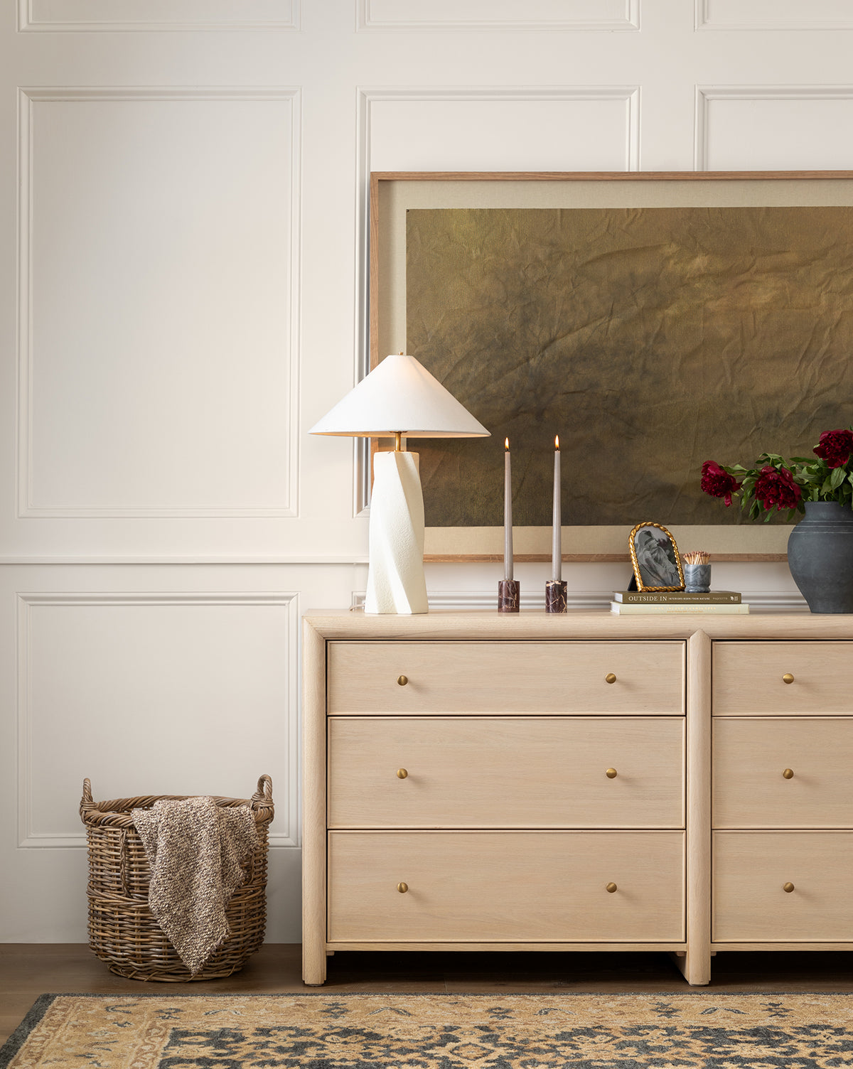 A light wood dresser with gold knobs stands against a white paneled wall, topped with a modern white lamp, candles, red flowers in a vase, and books. A Mckell Basket and framed art add to the cozy decor of natural materials.