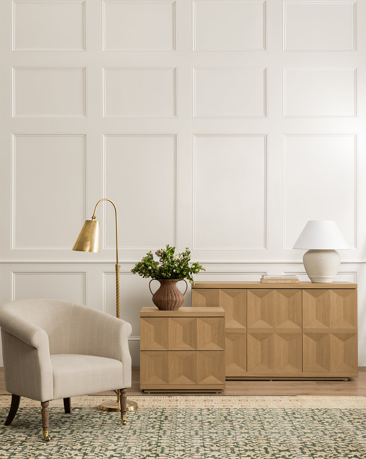 A light beige armchair sits next to a brass floor lamp by a white paneled wall. Two Beacon Nightstands with geometric patterns provide modern storage for books and plants, while a patterned rug completes the look, by McGee & Co.