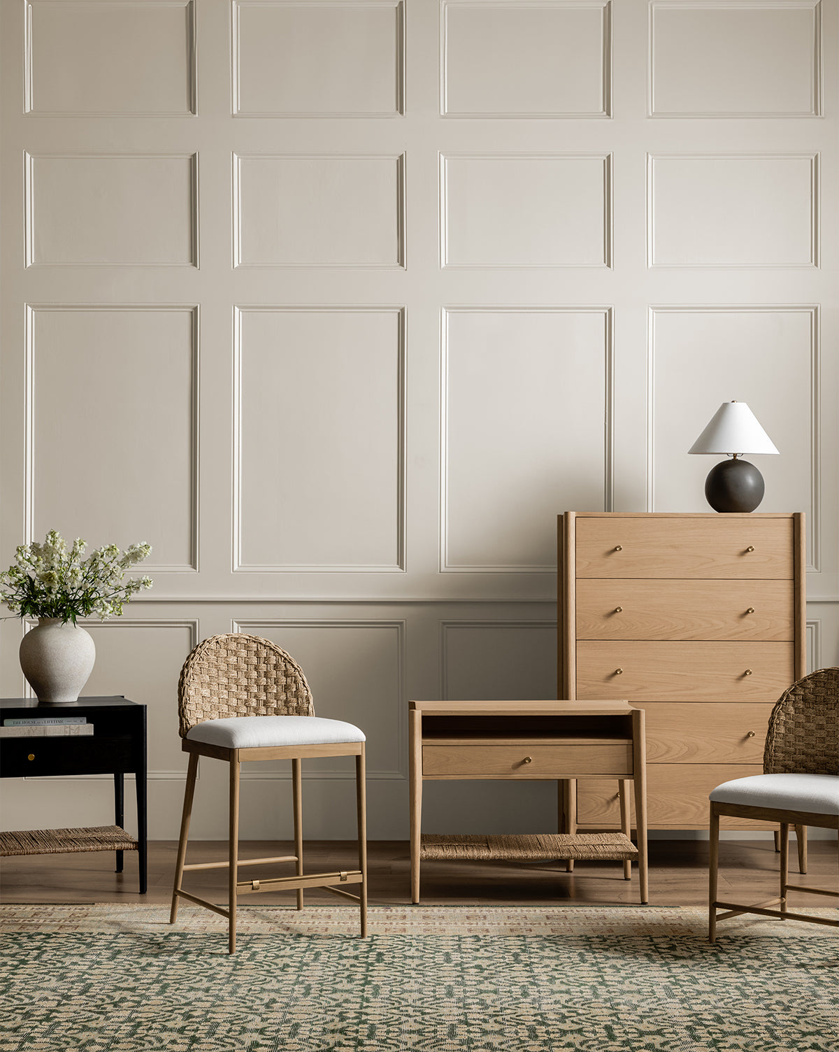 A stylish room with a cream paneled wall, Dana White Oak Nightstand with brass hardware, two woven-back chairs, black side table with white flowers, small lamp, and a green patterned rug by McGee & Co.
