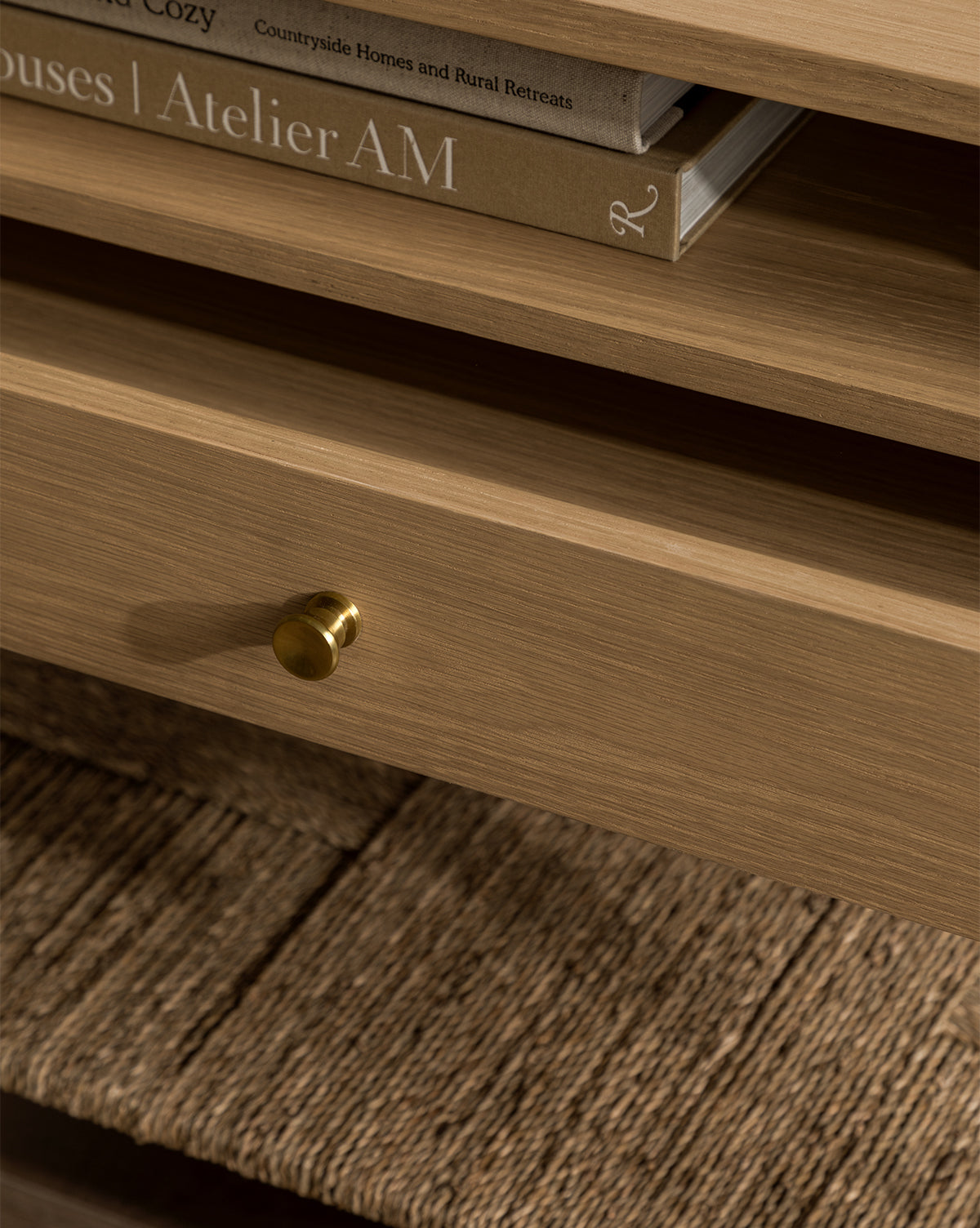 Close-up of the Dana White Oak Nightstand featuring a small brass knob, an open shelf with two stacked books, and a textured woven seagrass shelf below the drawer, by McGee & Co.