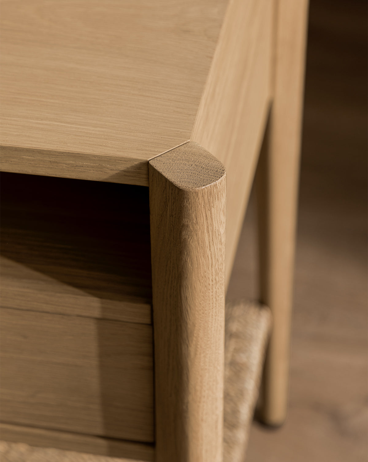Close-up of the Dana White Oak Nightstand corner featuring smooth, rounded edges and visible natural wood grain. A woven seagrass shelf appears below, with the light finish showcasing its finely crafted details by McGee & Co.