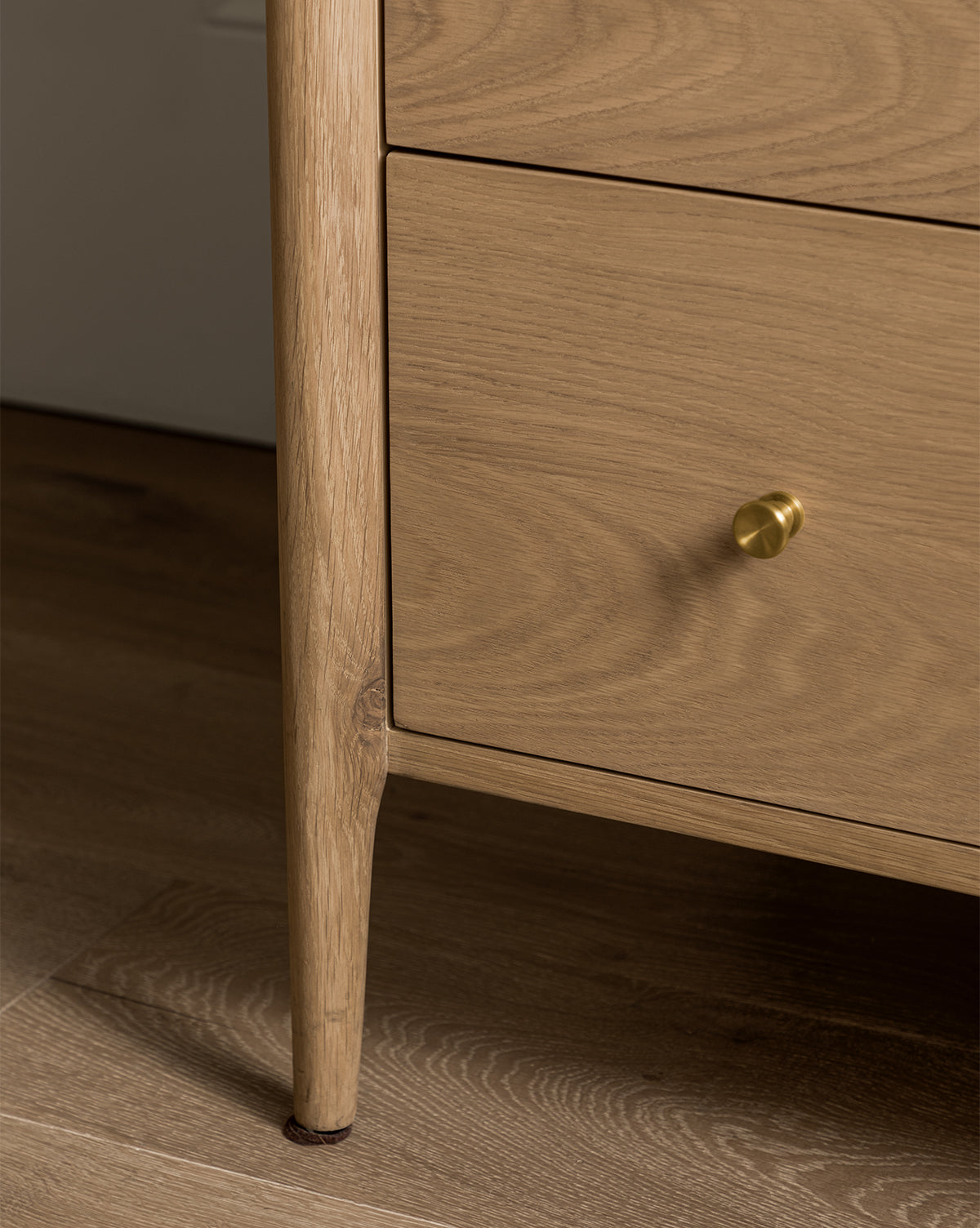 Close-up of the Dana Tallboy Dresser’s lower corner, featuring white oak with light wood grain, a round brass knob, and a tapered leg standing on a wooden floor, by McGee & Co.