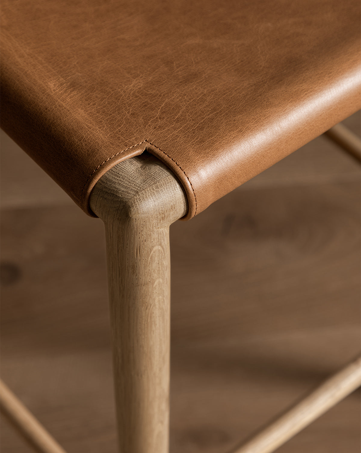 A close-up of the Danson Counter Stool corner shows its smooth leather seat with visible stitching, neatly wrapped over a rounded light wood frame. The stool’s minimalist design pairs seamlessly with a similarly toned wooden floor in the background, by McGee & Co.