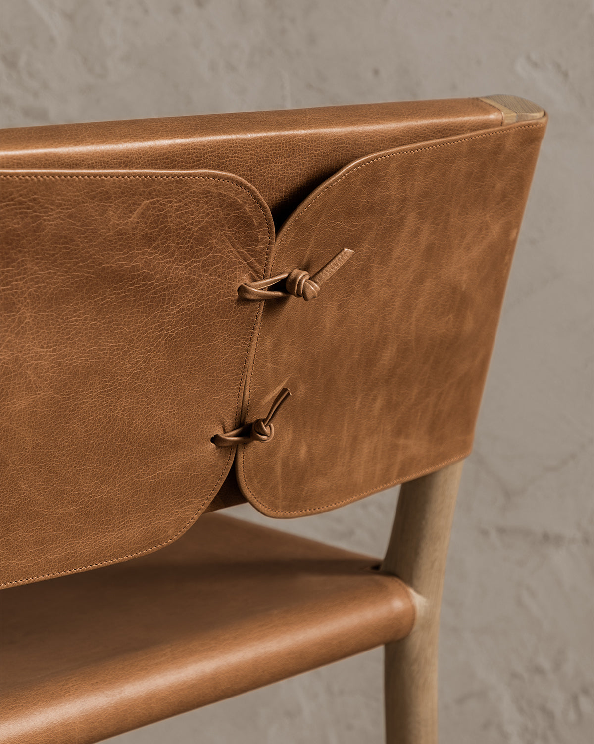 Close-up of the Danson Counter Stool, showcasing its light brown leather seat and backrest with two panels connected by leather cords, highlighting minimalist design against a light textured wall, by McGee & Co.
