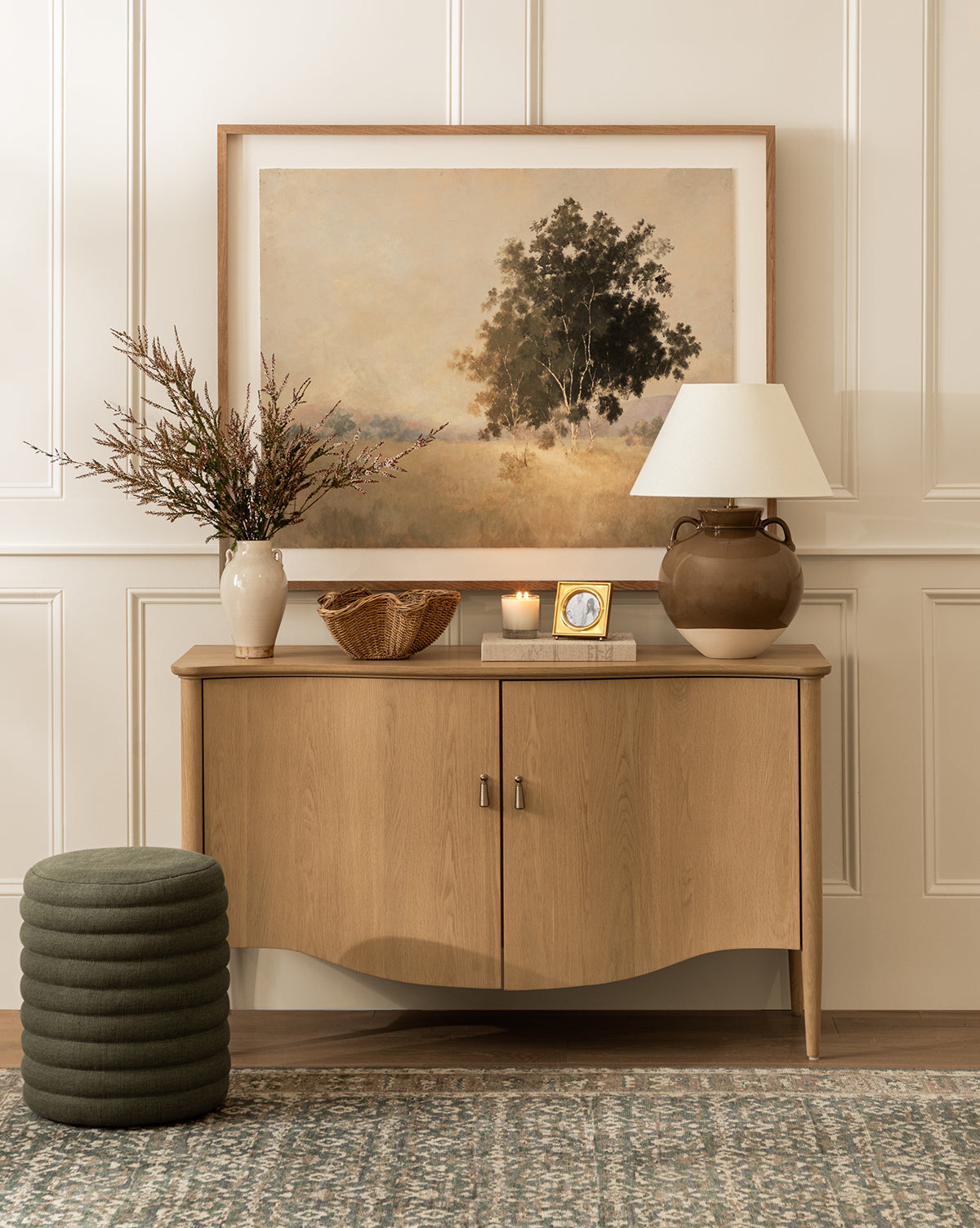 A wooden console table displays We Woke To A Dream by Aileen Fitzgerald in a white oak frame, a lamp, vase with branches, candle, clock, and bowl against a white paneled wall; in front are a green ottoman and patterned rug by McGee & Co.