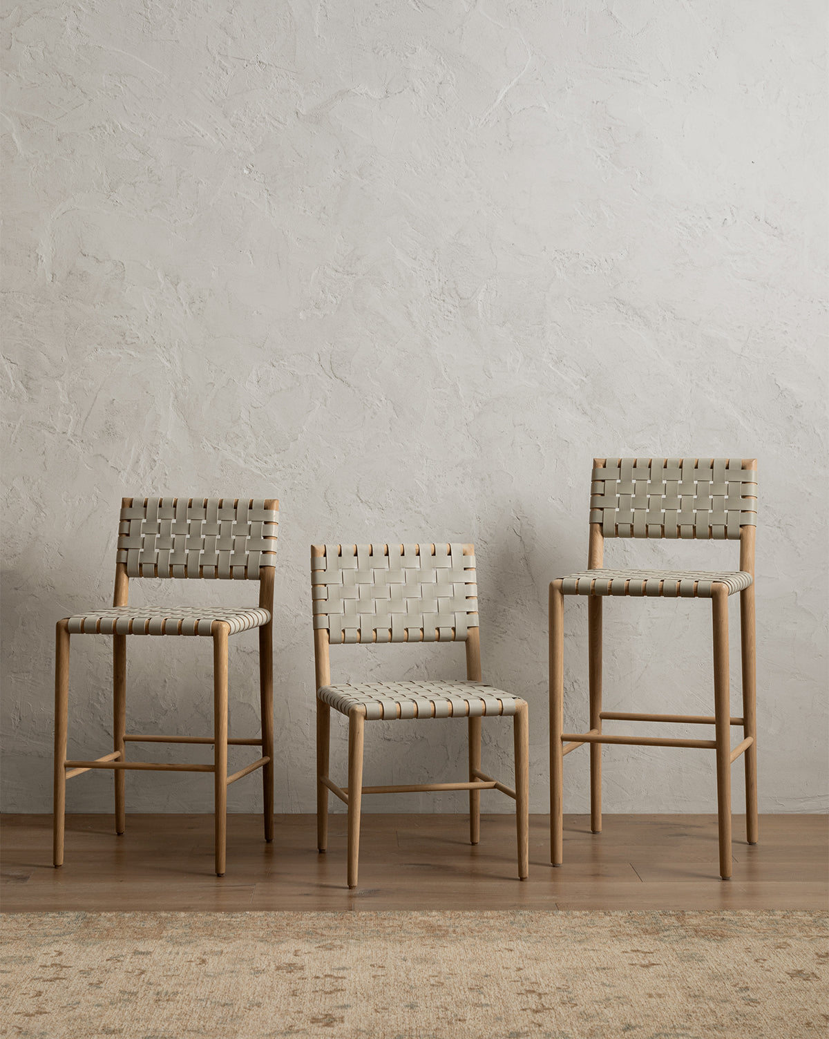 A Greely Dining Chair and two tall tan wooden barstools with woven backs are arranged side by side against a textured light gray wall on a wooden floor, by McGee & Co.