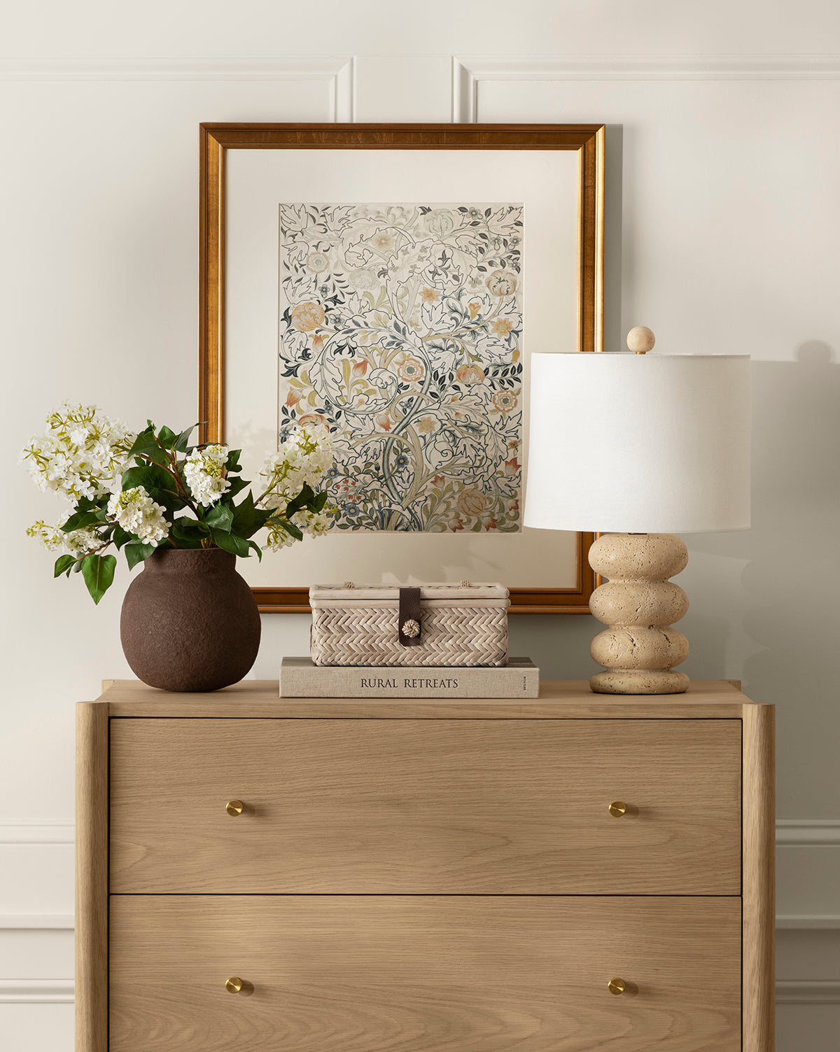 A light wood dresser with two drawers displays a brown vase of white flowers, books, a patterned box, and a beige lamp. Above, the Acanthus PortieÌ€re botanical wall art in an antique gold frame hangs on the white paneled wall.