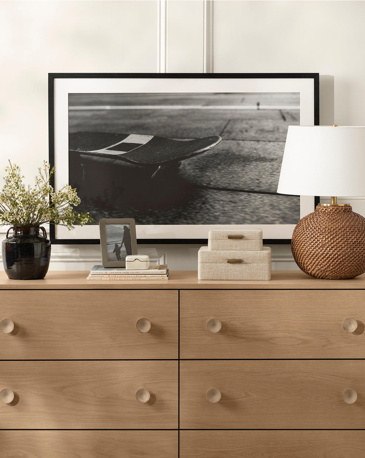 A light wood dresser with round knobs displays the Avedon Table Lamp with a white linen shade, two fabric boxes, a black vase of flowers, a photo frame, and books. Behind them rests a framed black-and-white skateboard photo by McGee & Co.