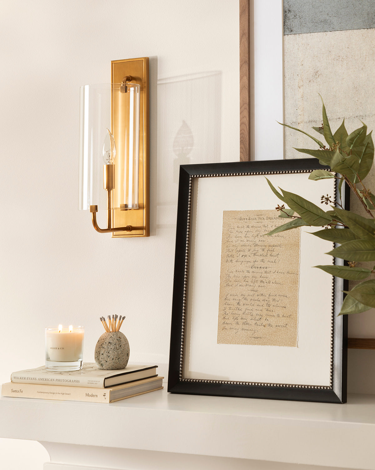 A gold wall sconce with a glass shade is mounted over a mantel featuring a candle, books, a pencil holder, and Give Back the Dreams, gallery-quality framed handwritten song lyrics, beside a leafy green plant.