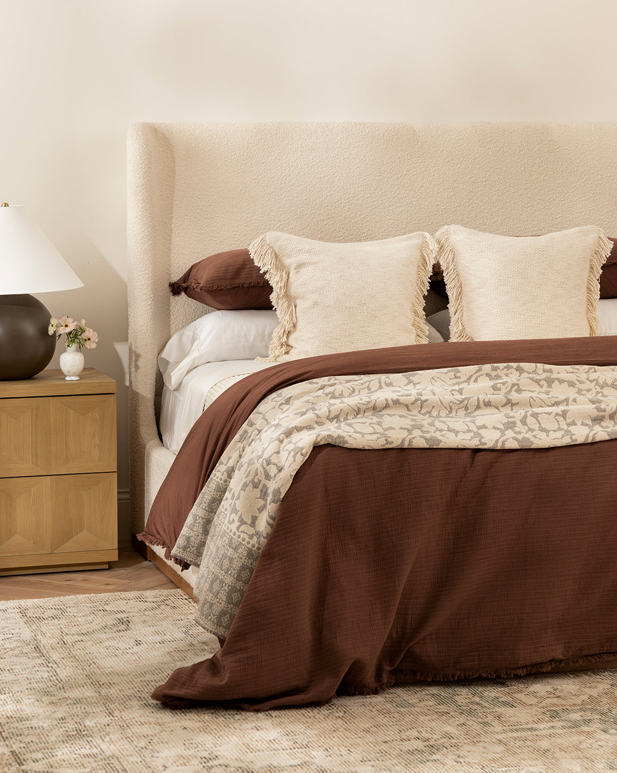 A cozy bedroom with a beige upholstered headboard, cream and brown fringe pillows, floral comforter, soft bedding topped by the Hendricks Chocolate Cotton Duvet Cover. A wooden nightstand holds a white lamp and flowers on a patterned rug.