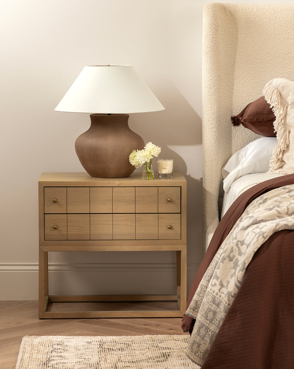 A wooden bedside table holds the Parma Table Lamp, featuring an ivory linen shade and textured ceramic base, next to a glass with white flowers and a candle. It stands beside a bed with brown bedding, a textured headboard, and part of a rug on the wood floor by McGee & Co.