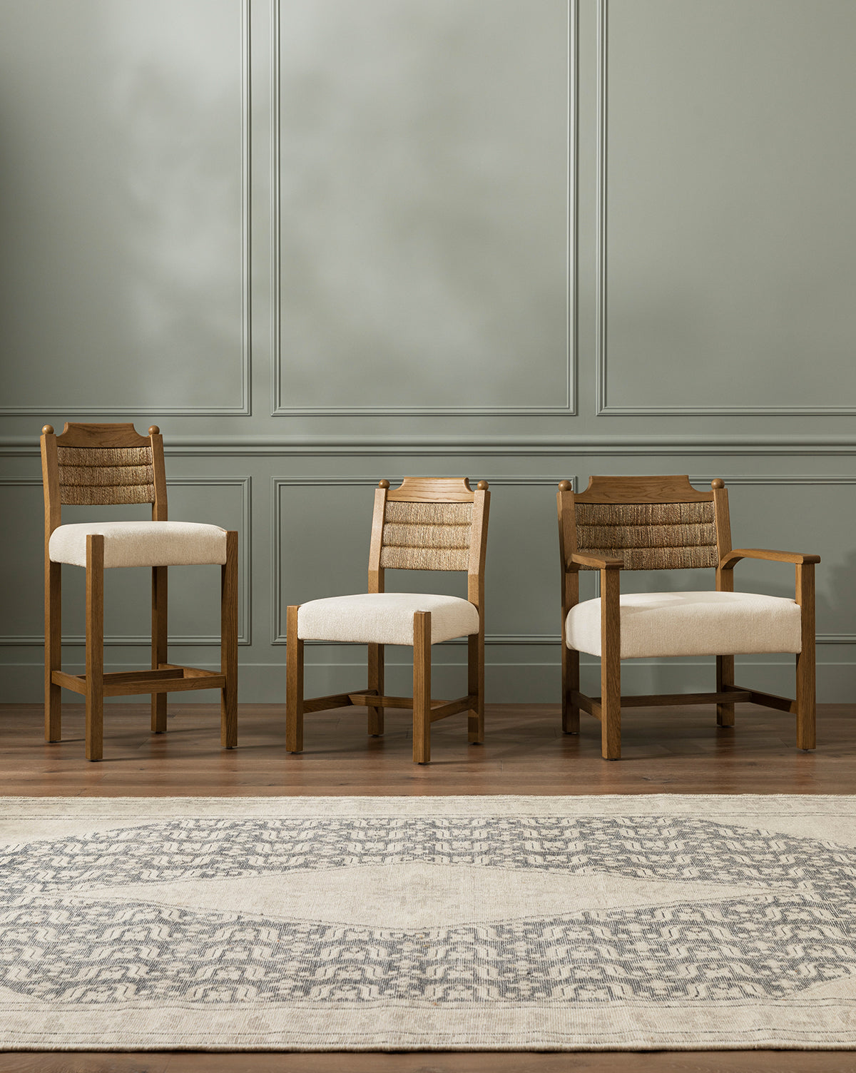 Three Alberta Dining Chairs with beige cushions and woven rattan backs are side by side on a wooden floor against a light gray paneled wall, with a patterned area rug in the foreground, by McGee & Co.