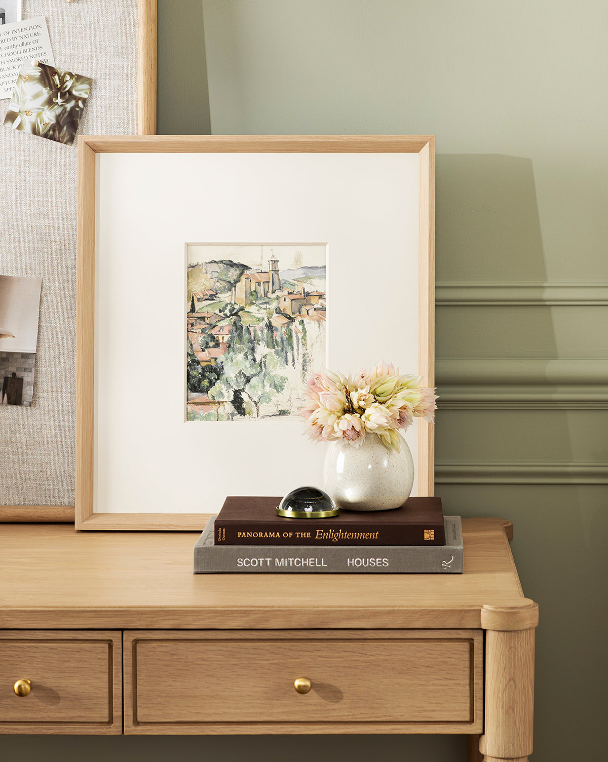 A wooden desk with two drawers displays Le Village, a museum-quality giclee print in a natural woodgrain frame, alongside pale flowers in a vase, decor objects, two stacked books—Enlightenment and Houses—and a partially visible pinboard by McGee & Co.