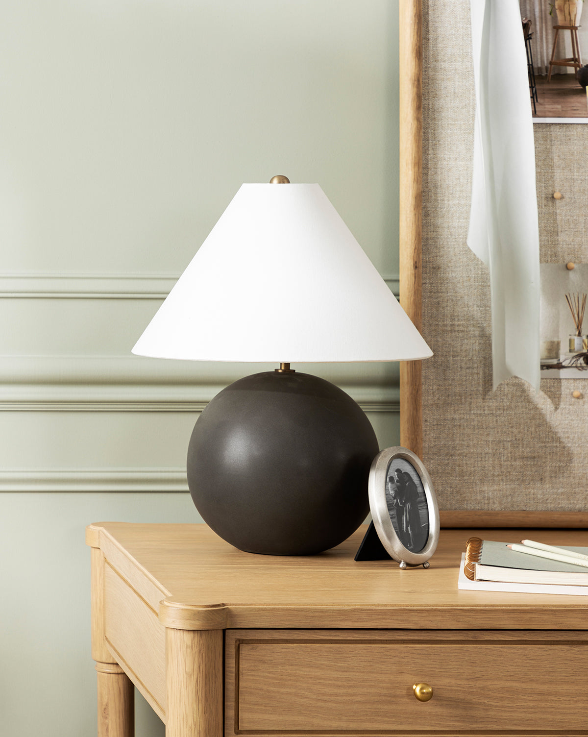 A wooden desk with a drawer holds a round black lamp with a white shade, the Blanche Pewter Picture Frame, some books, and a notepad. Light green walls with molding and a fabric-covered pinboard are in the background.