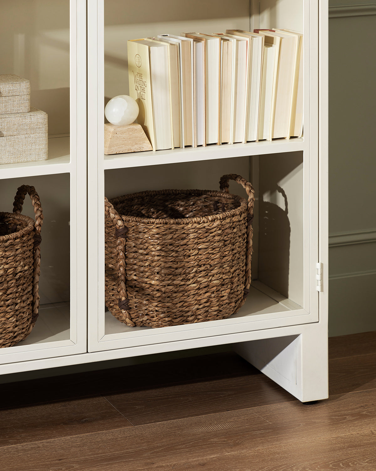A white bookshelf holds cream books on the top shelf, Salter Woven Baskets on the lower shelf, and a small lamp with a round bulb beside stacked books, all arranged on a wood floor by McGee & Co.