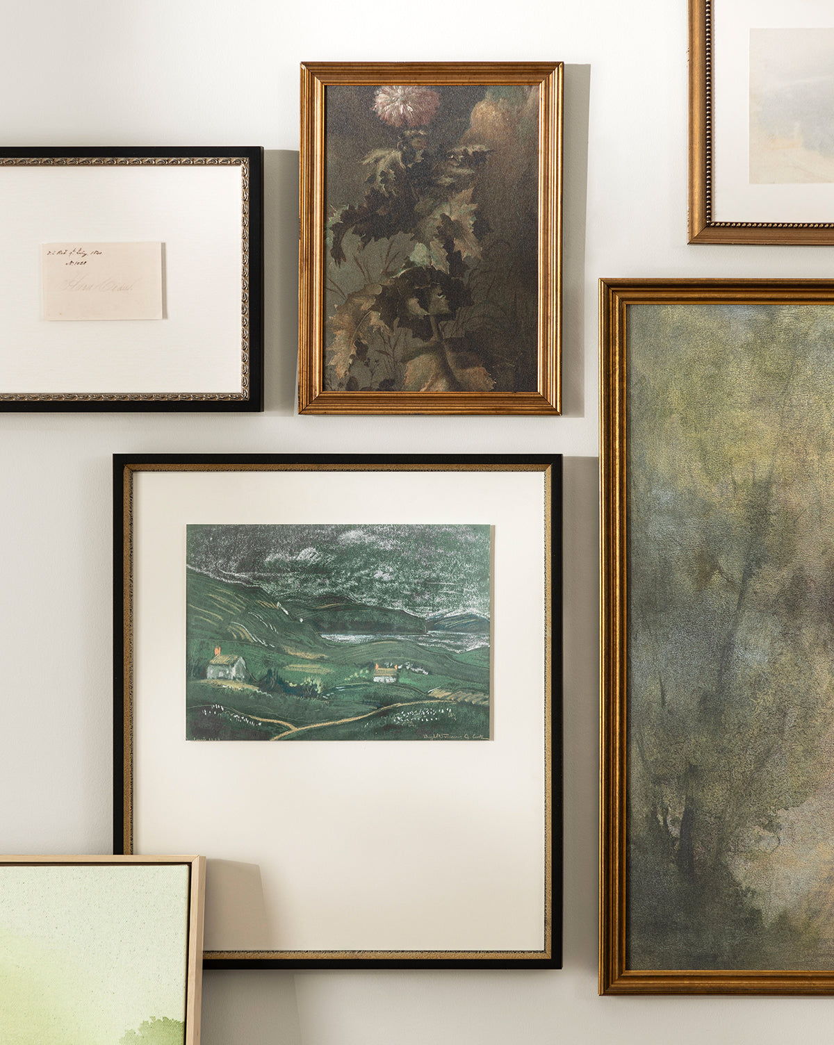A gallery wall displays various framed artworks, including Thistle Down, an abstract painting, a green hills landscape, flower wall art in an antique gold frame, and a handwritten note—all beautifully arranged on a light wall by McGee & Co.